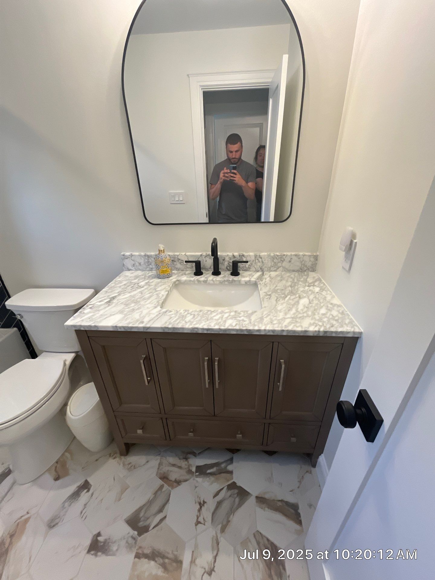 Bathroom with brown vanity, white countertop, black faucet, and unique mirror. Toilet to the left and person in mirror.