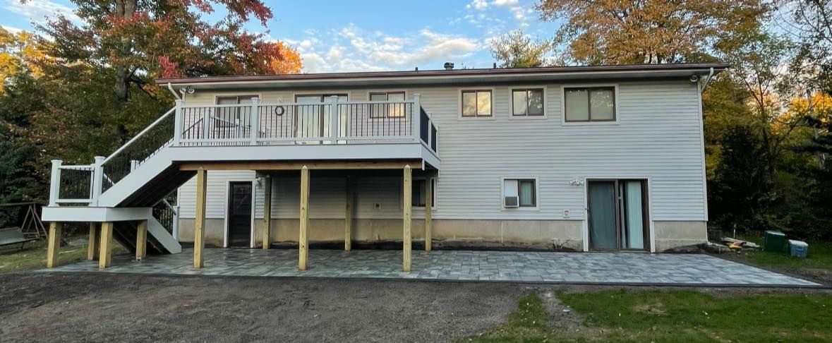 A two-story house with a deck and patio, surrounded by trees and a blue sky.