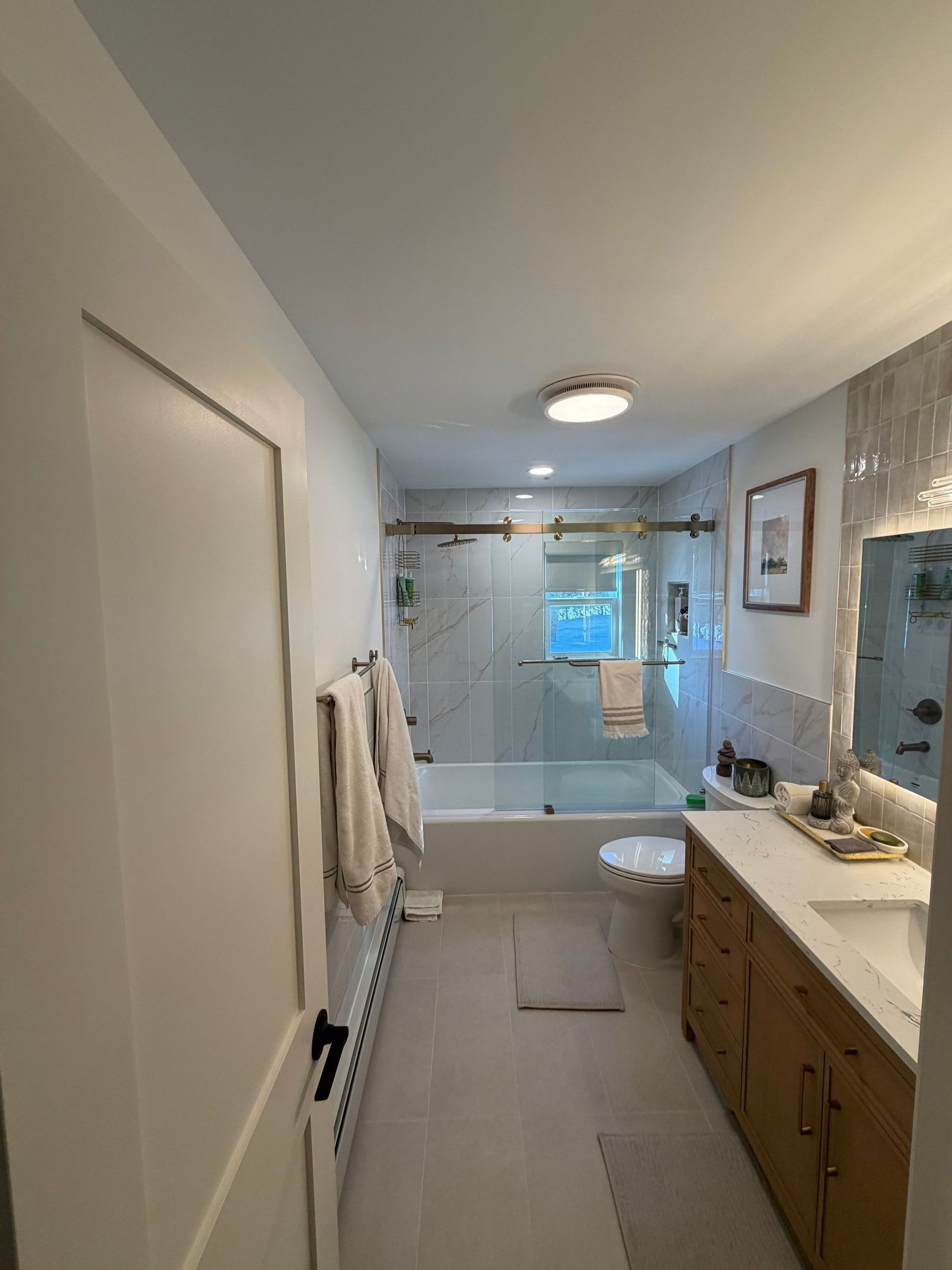 Bathroom with a tub and shower, toilet, and vanity. White walls and fixtures, wood vanity, and a clear glass shower door.