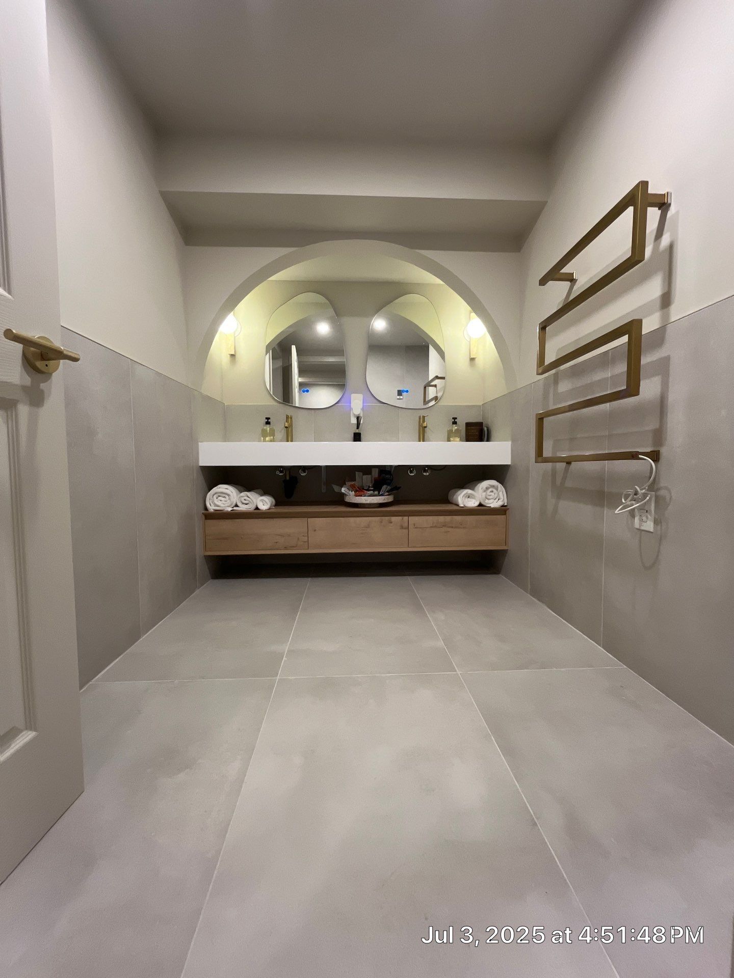 Bathroom with arched mirrors above a wooden vanity. Grey tiled floor, gold accents, and a towel warmer.