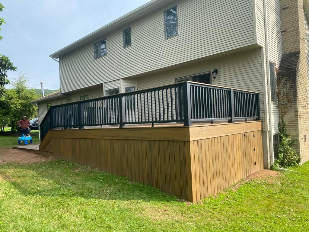 Two-story house with a wooden deck. Green grass and black railing, brown siding. Person nearby.