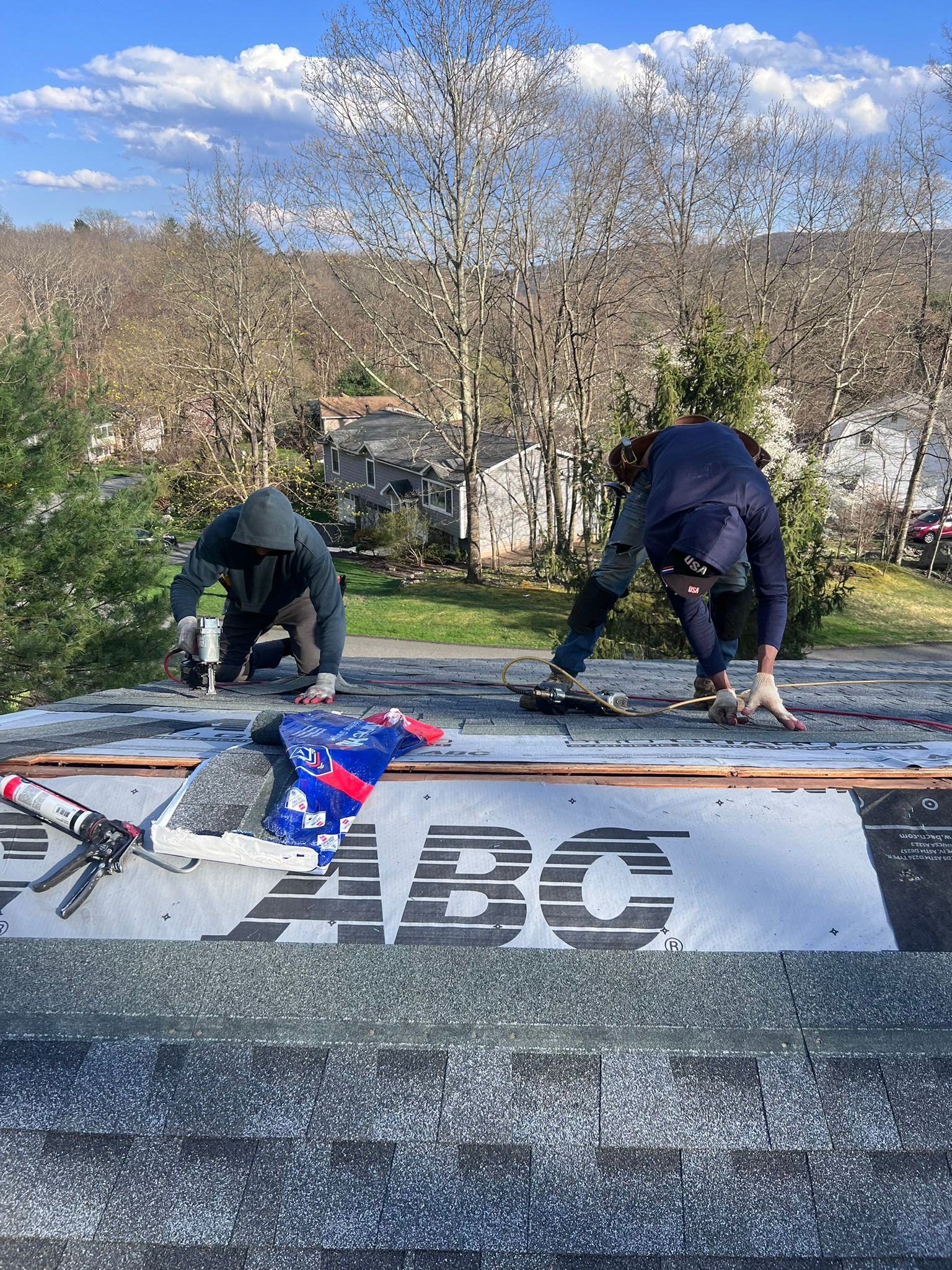 Two roofers on a rooftop installing shingles. The roofers are using tools in bright sunlight.