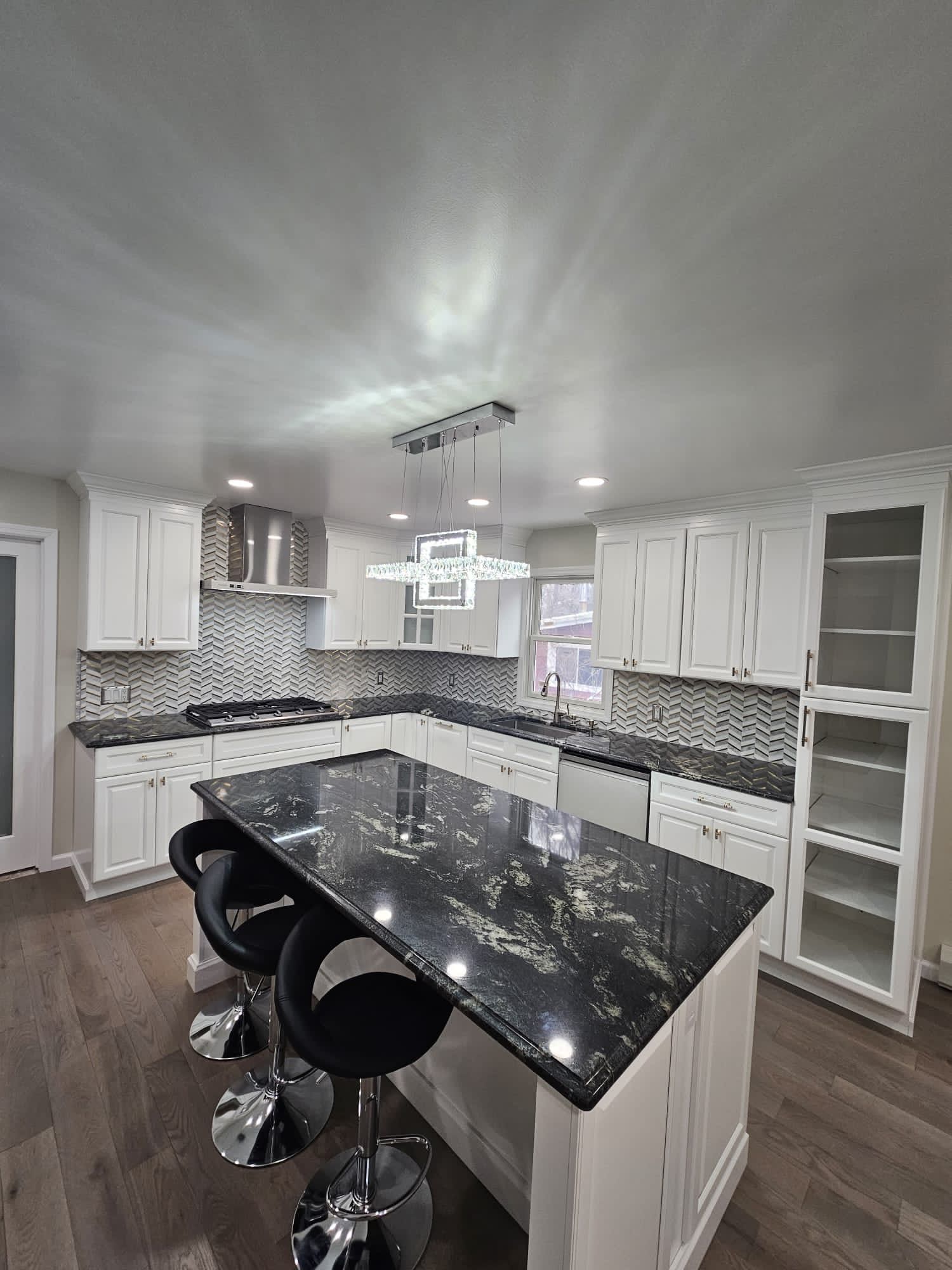Modern white kitchen with black countertop island, chrome barstools, stainless steel appliances, and mosaic backsplash.