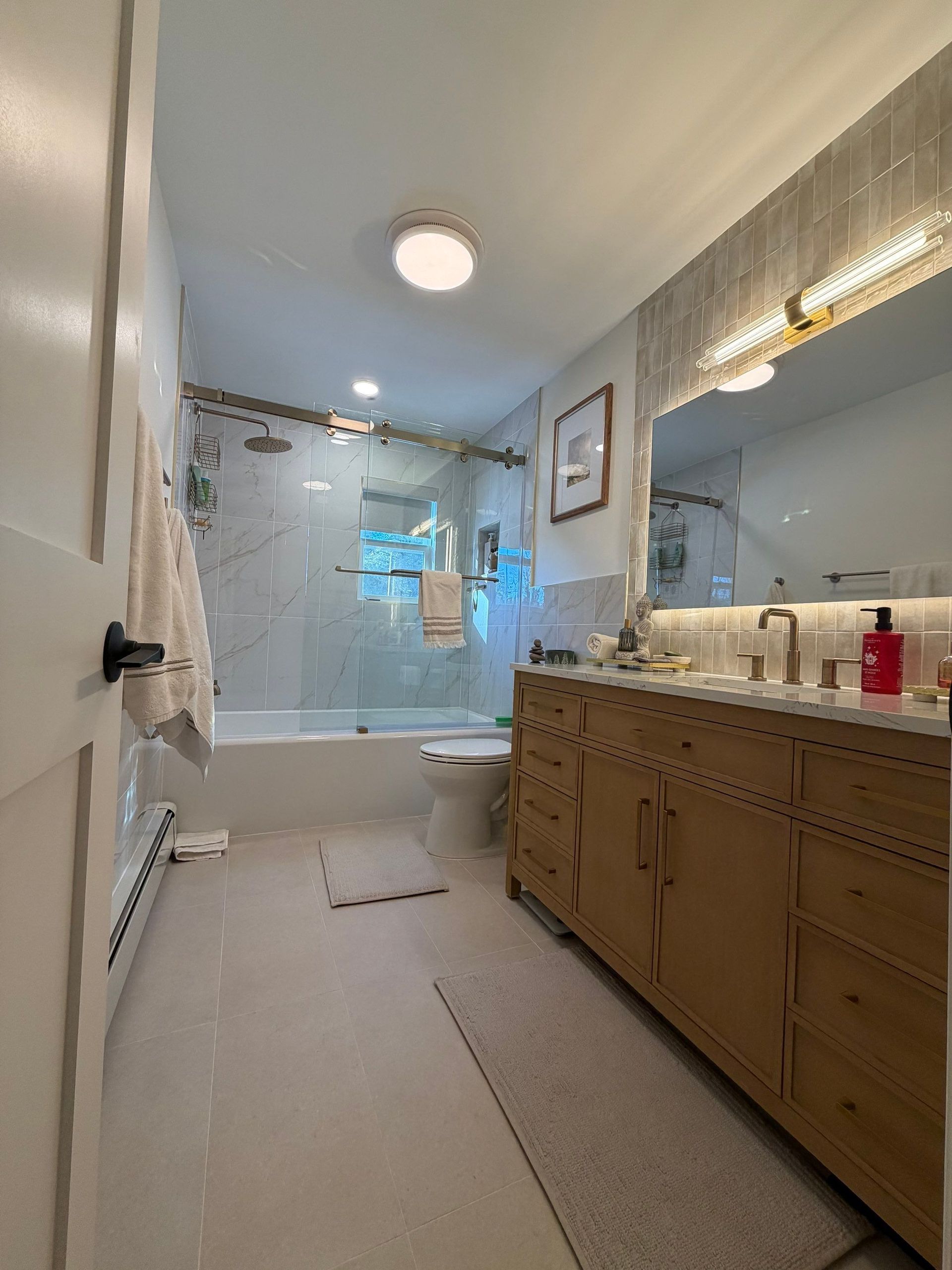Bathroom with a light wooden vanity, marble-tiled wall, glass shower door, and a white bathtub.