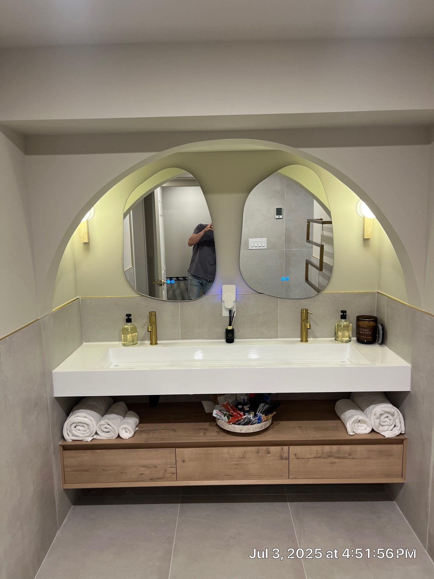 Modern bathroom with dual sinks, floating wood vanity, and two irregularly shaped mirrors under an arch.
