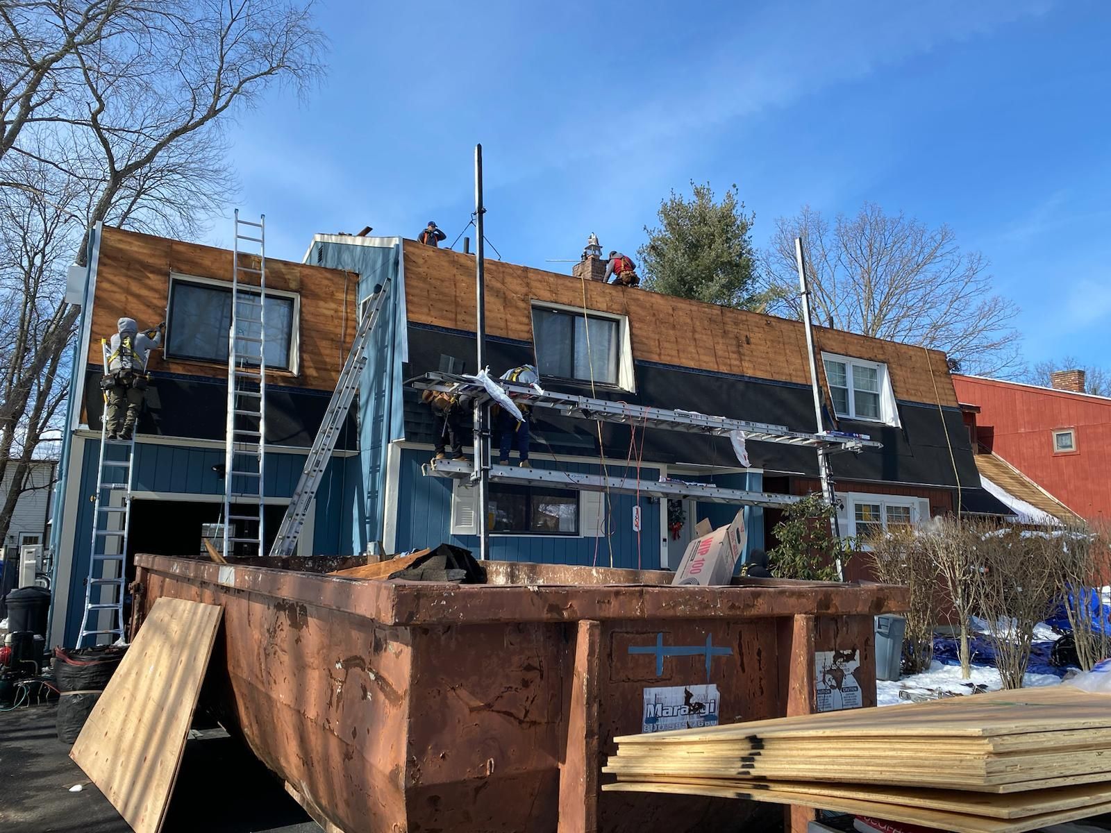 Workers on a roof replacing shingles, blue building, brown dumpster, ladder, clear sky.