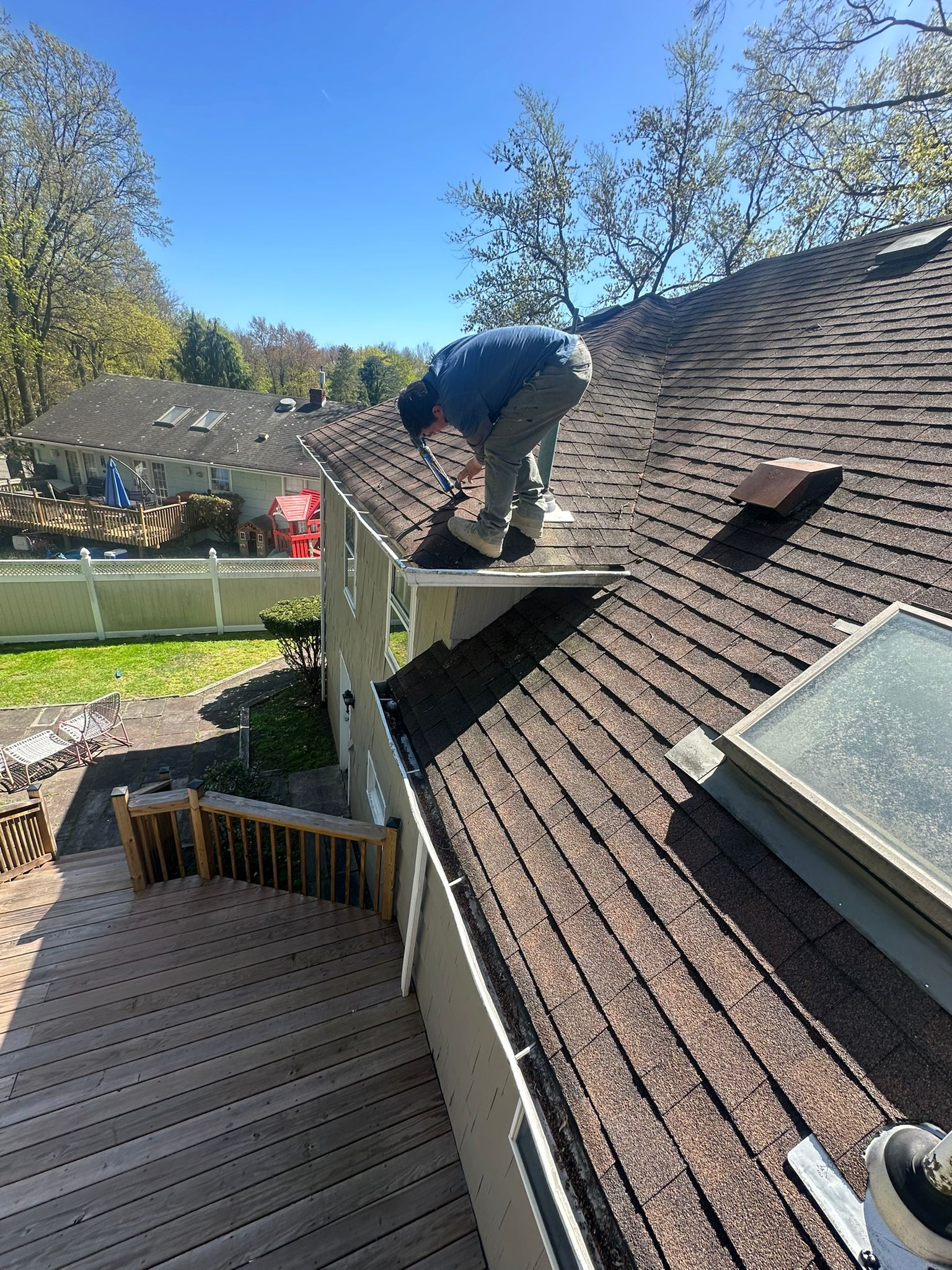 Person on a roof repairing shingles on a sunny day. Brown roof with skylight, beige house, and wooden deck.