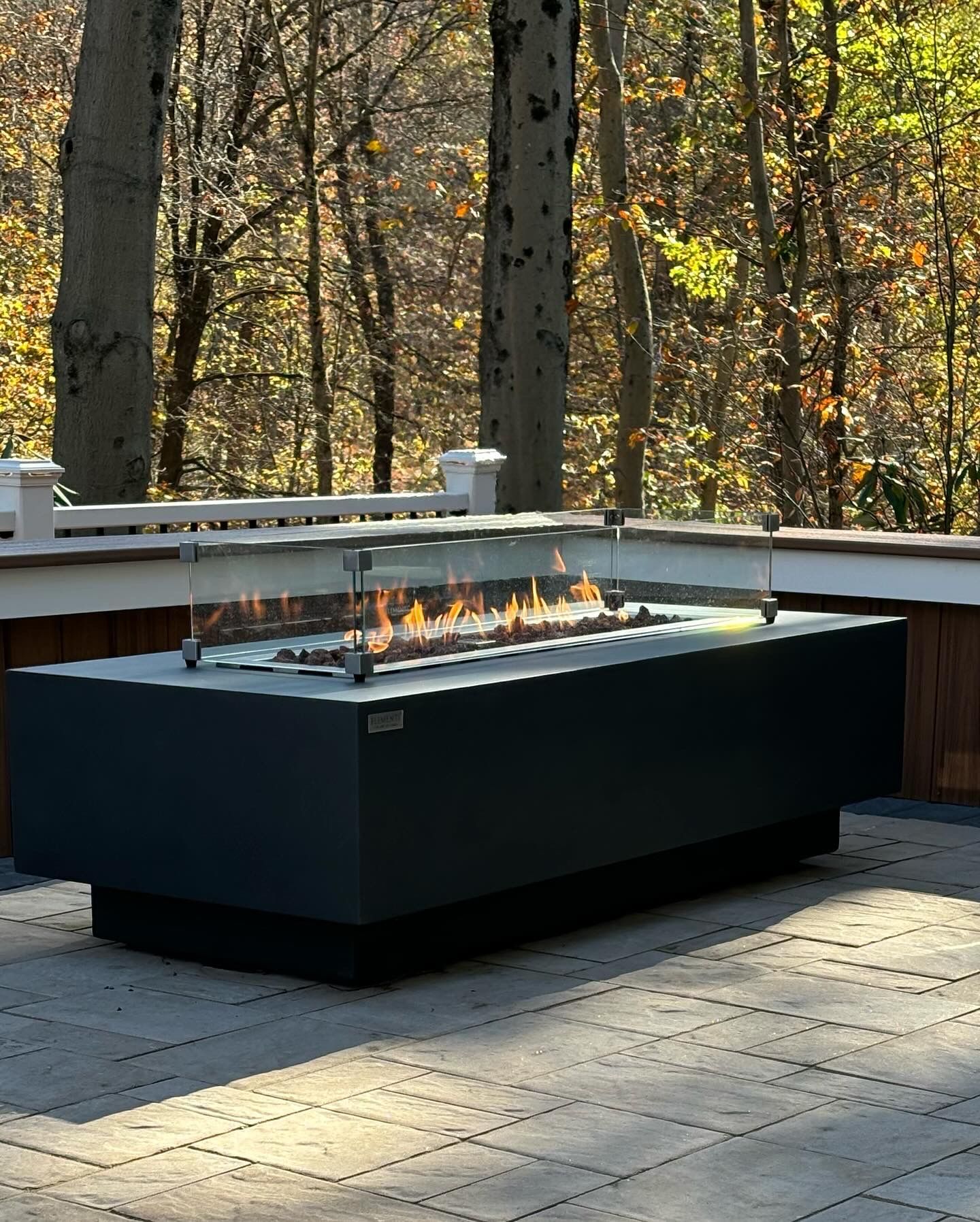 Modern rectangular fire pit on patio, surrounded by trees and wooden deck. Flames visible.