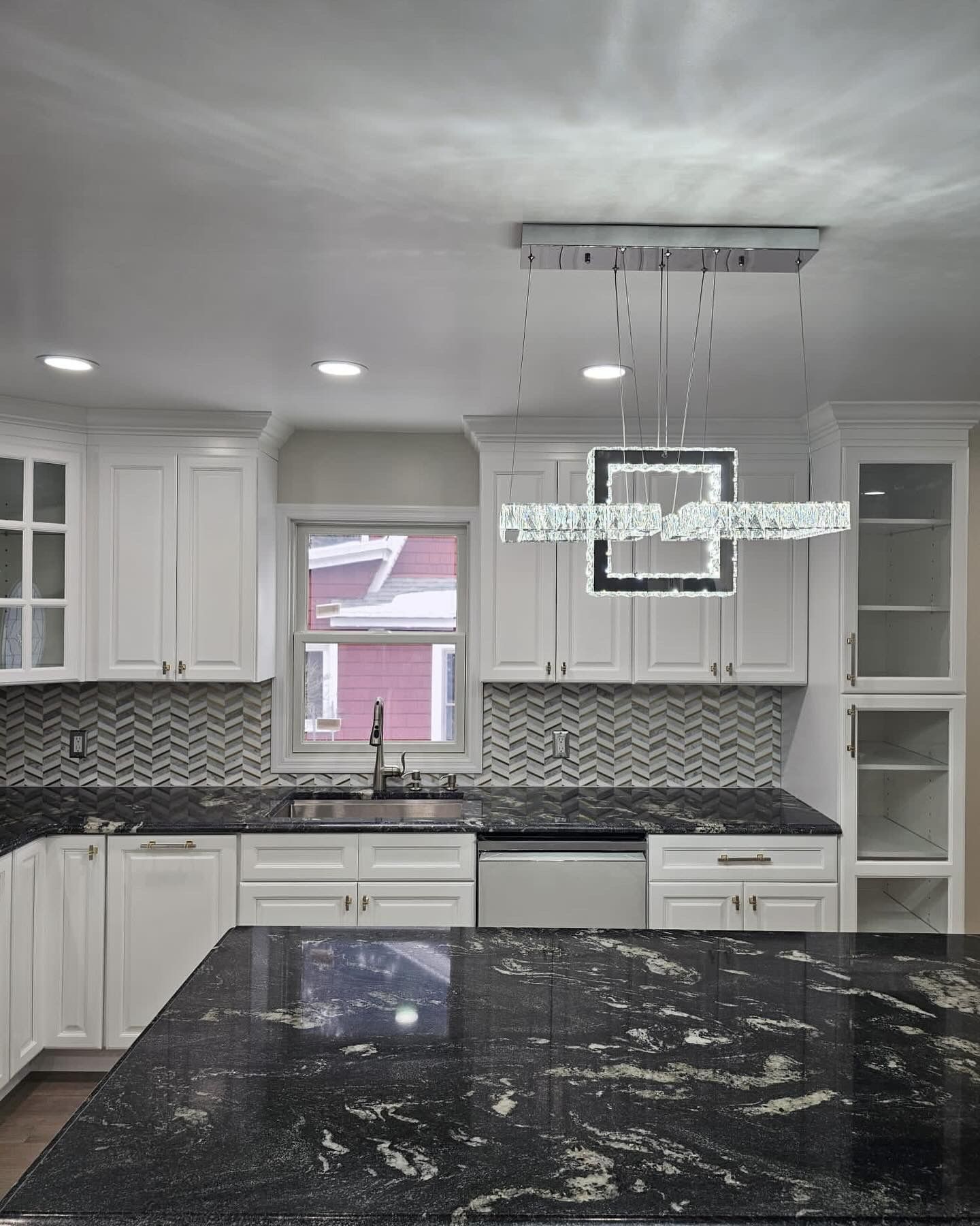 Modern white kitchen with dark granite countertops and decorative light fixture.