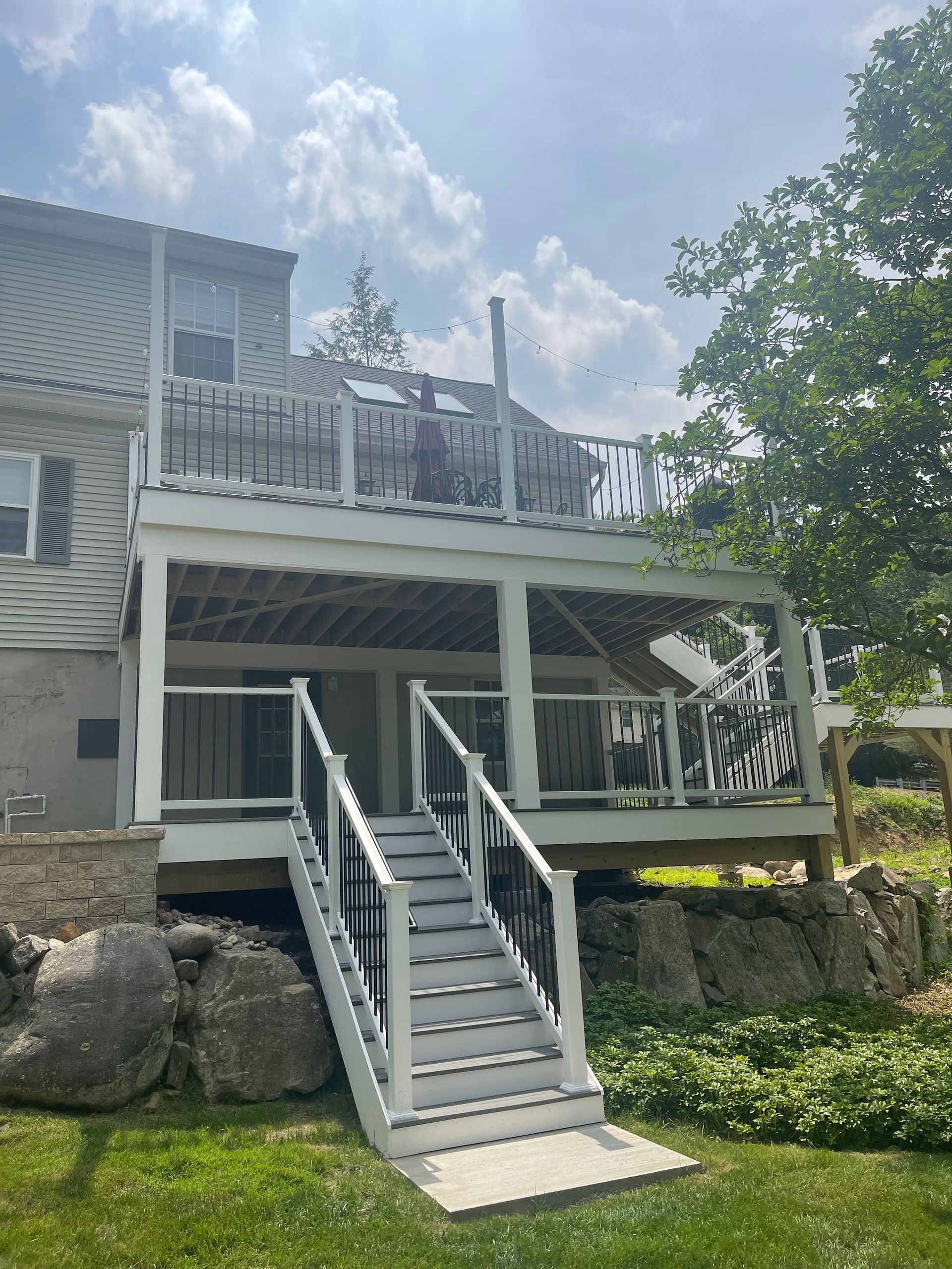 Two-story white deck with stairs leading down to a concrete pad. Gray house to the left, green grass and rocks below.