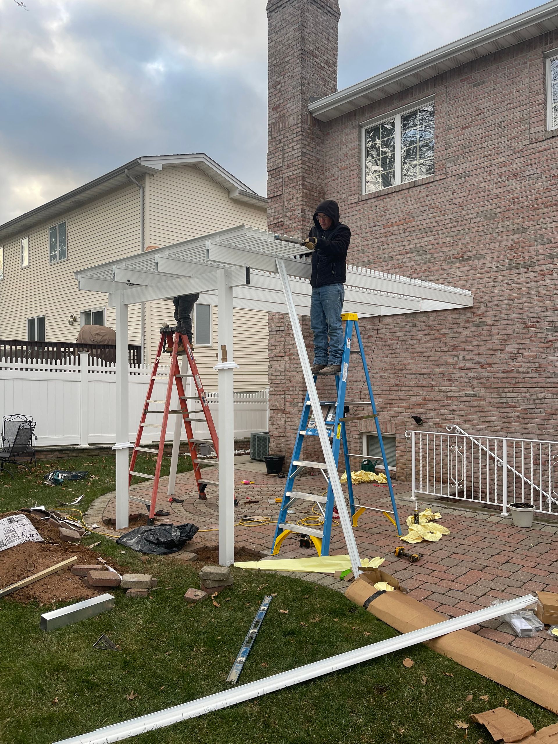 Construction workers building a white pergola on a brick patio next to a house with a chimney.