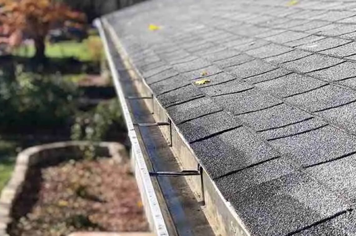 Close-up of a house roof with a gutter filled with leaves. Shingles are gray.