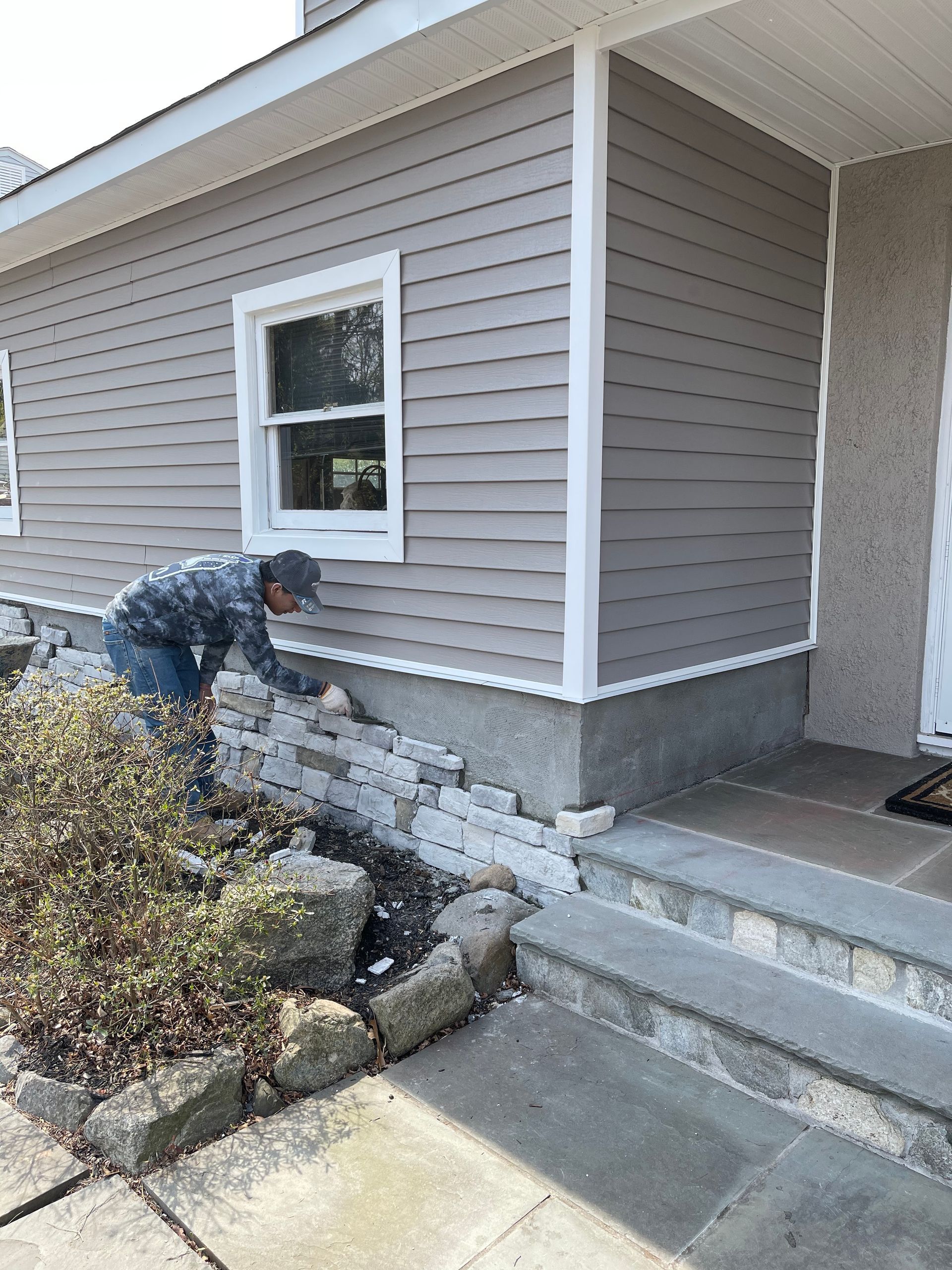 Person installing stone veneer on the exterior of a house; siding and window are above.