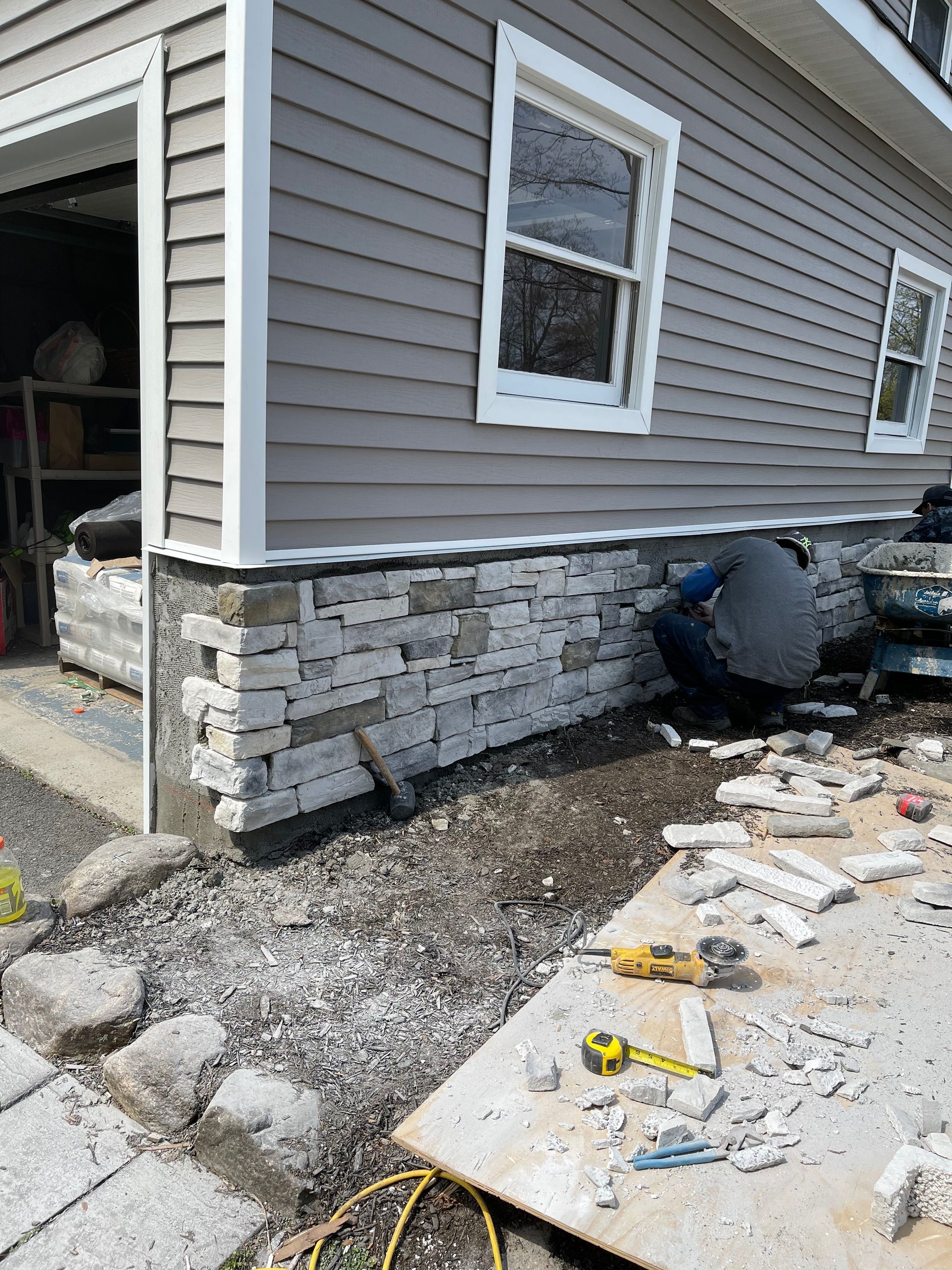 Construction worker installing stone veneer on the foundation of a gray-sided building next to an open garage door.