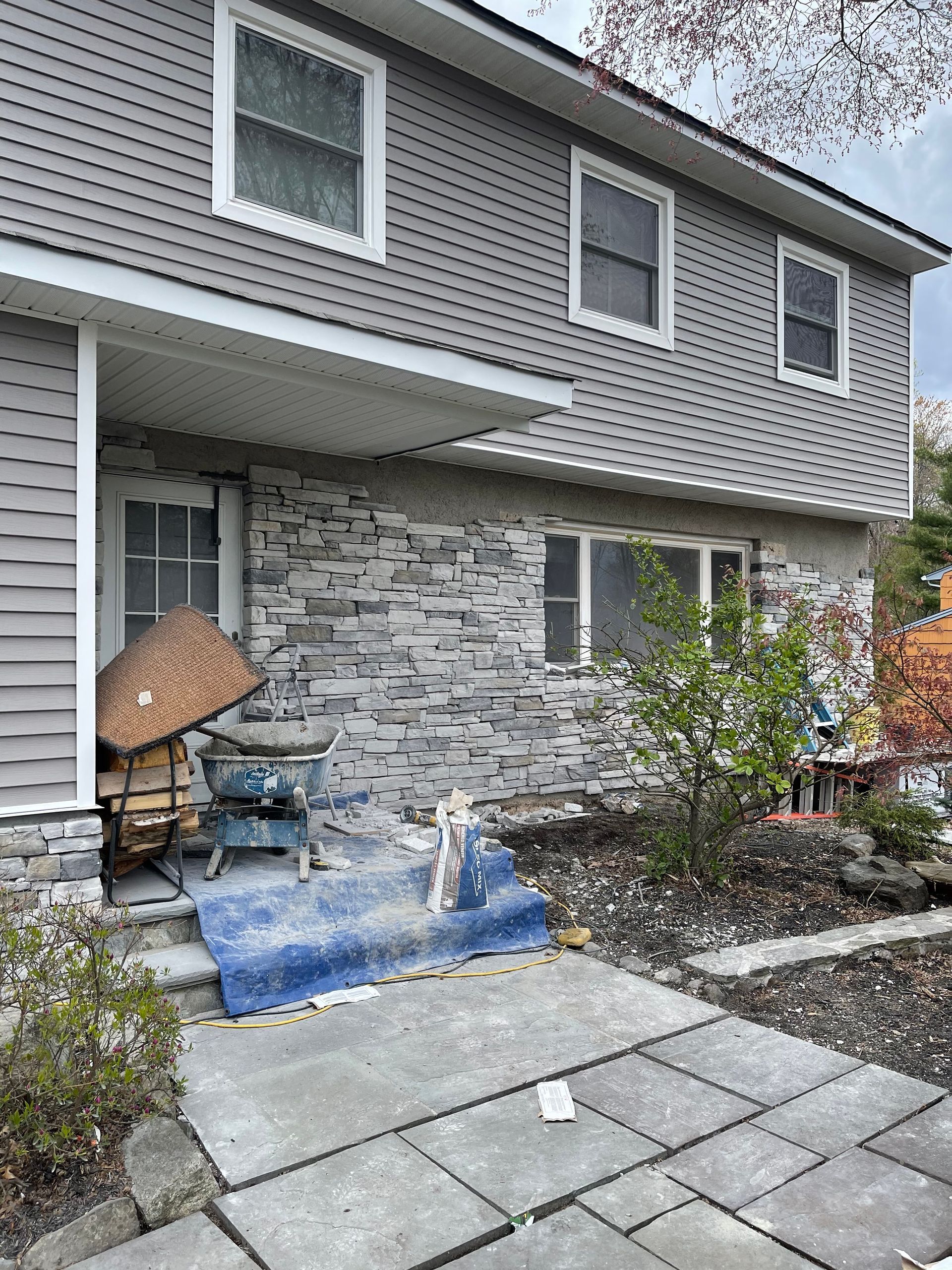 Two-story house with stone facade, gray siding, and a front entrance with a blue tarp and a wheelbarrow.