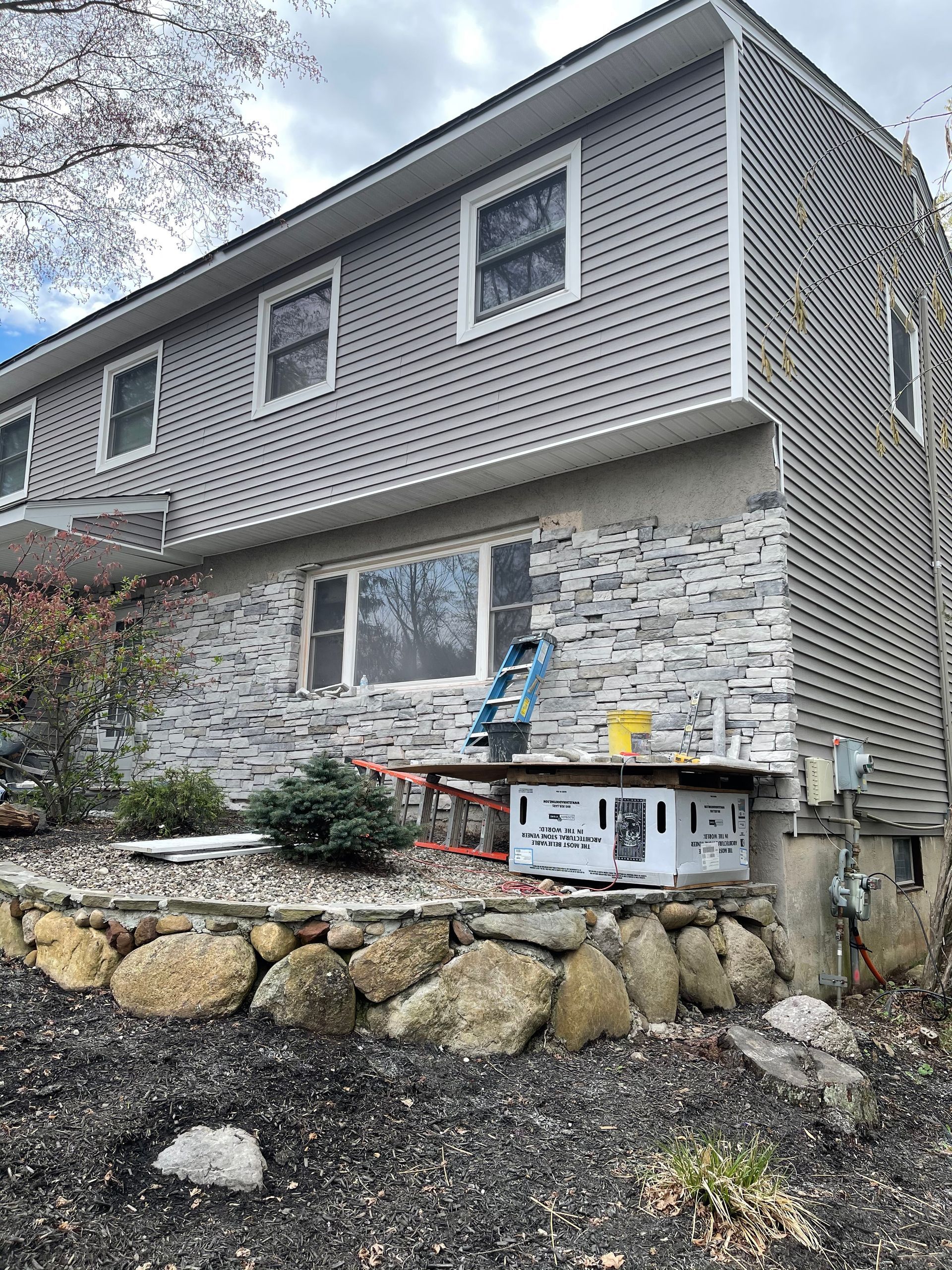 Two-story house with gray siding, stone facade, and construction equipment on a rock wall.