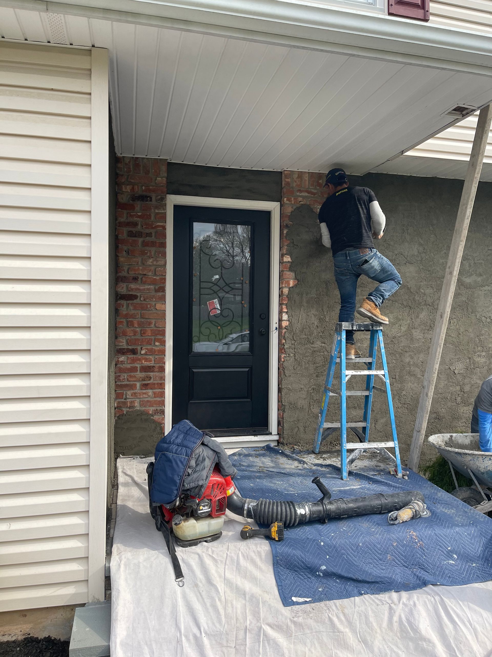 Man on ladder applying stucco to a house exterior near a black door. A blower and tarp are at the base.
