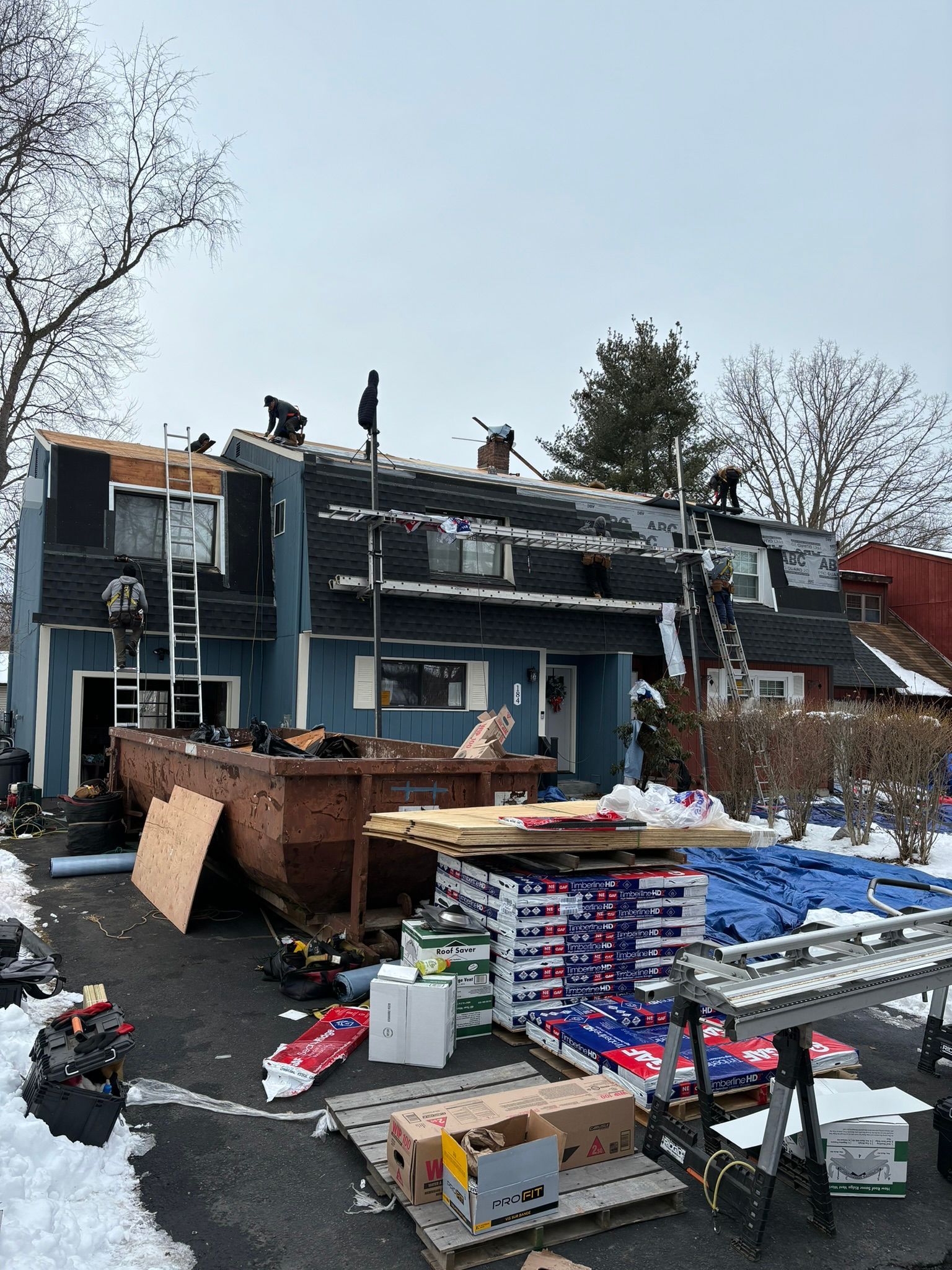 Roofing work in progress on a blue townhouse during winter. Workers, ladders, and supplies are present.