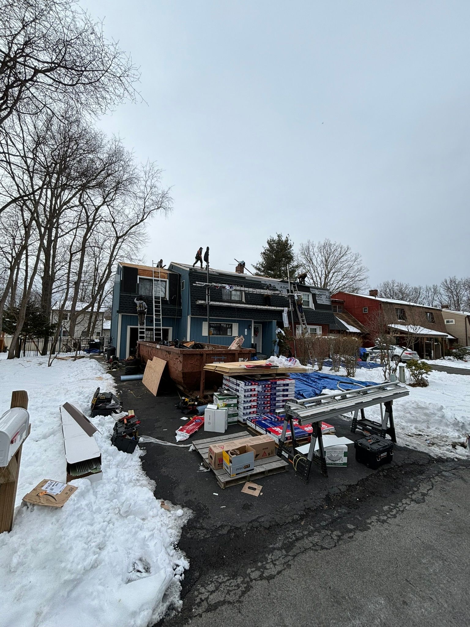 Two-story house with fire damage, debris in snowy yard. Gray sky, bare trees.