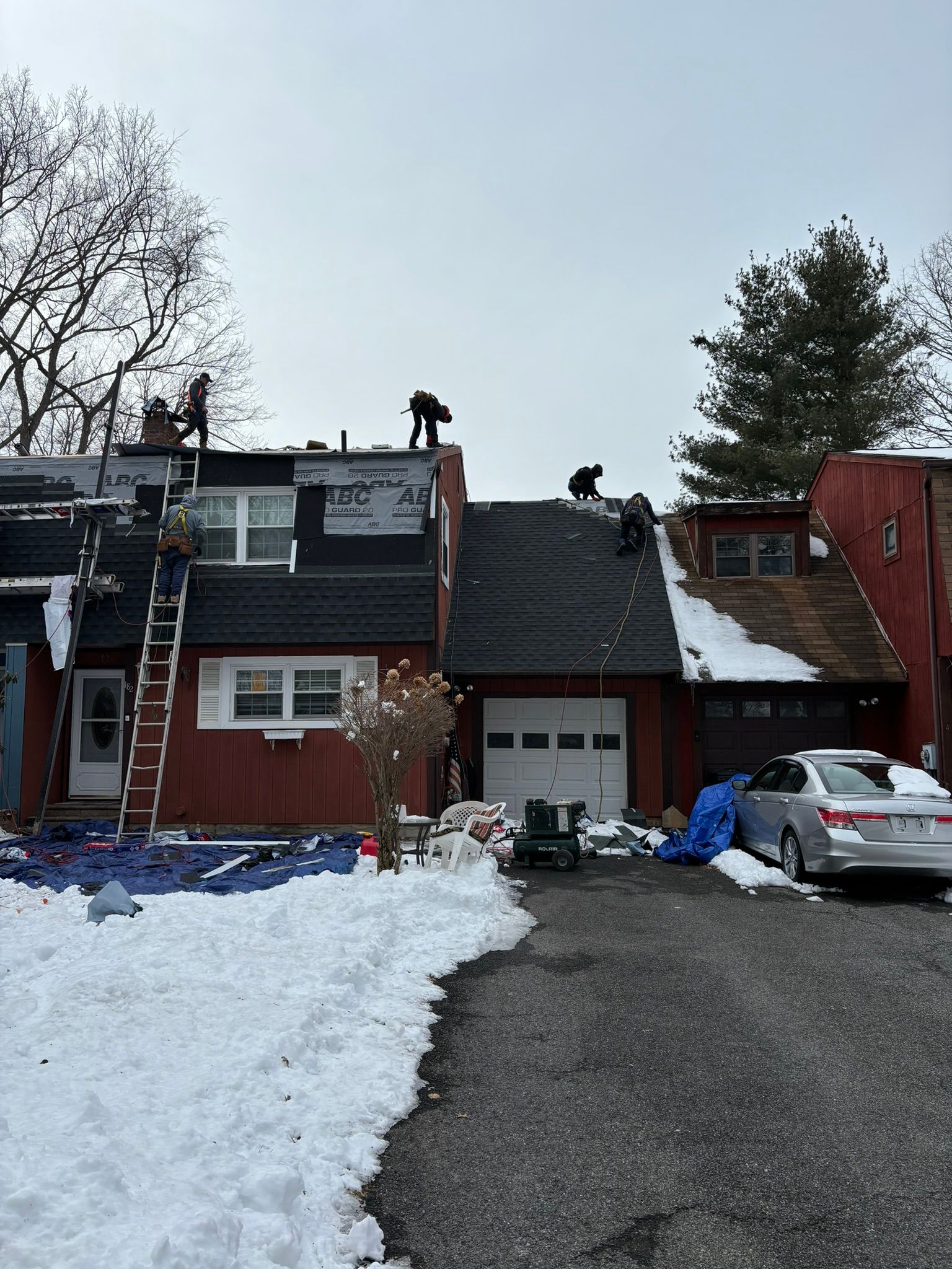 Roofers on a red townhouse roof in winter, installing shingles; snow on ground.
