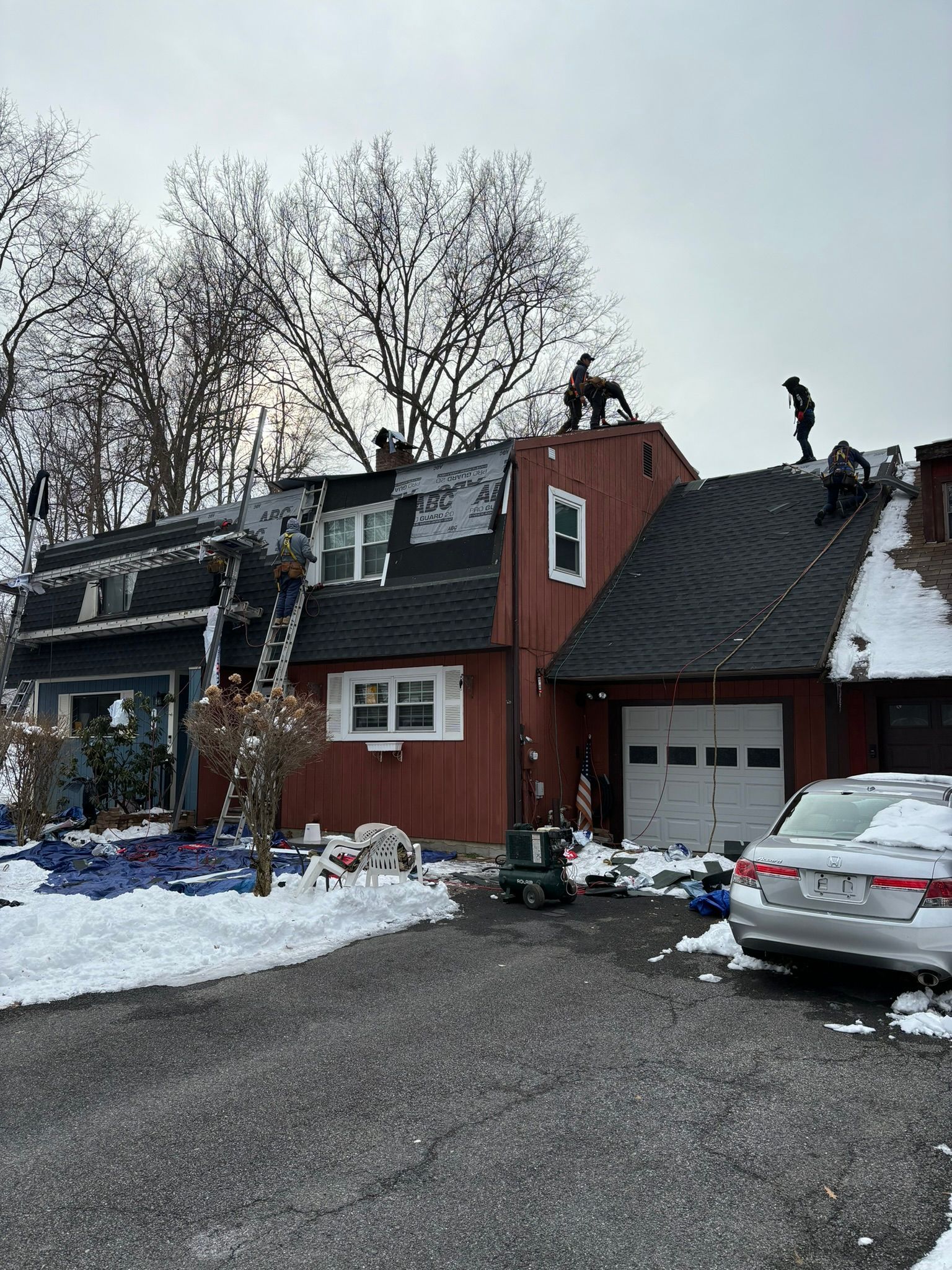 Roofers working on a snow-covered house with a white garage door. Trees and a car are in the yard.