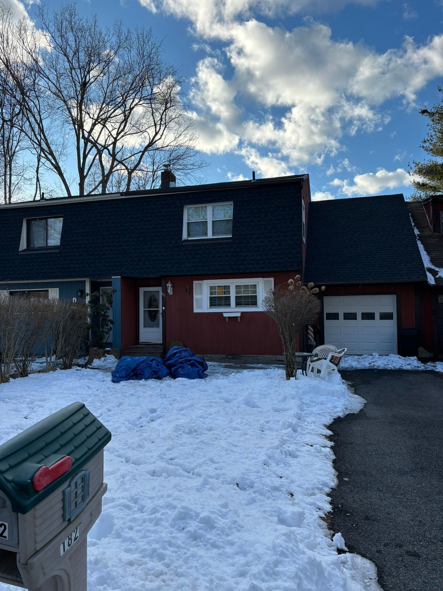 Snow-covered townhouse with red siding, dark roof, and a white garage door under a cloudy sky.