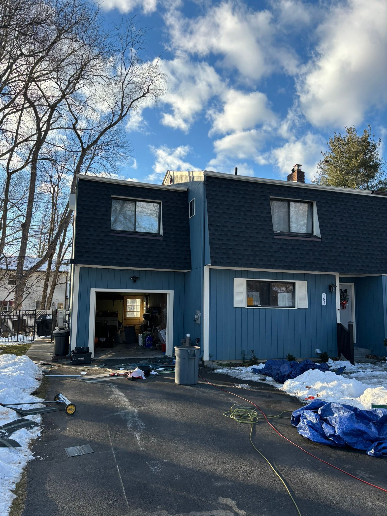 Blue townhouse under construction on a snowy day; tarp on driveway, bare trees, cloudy sky.