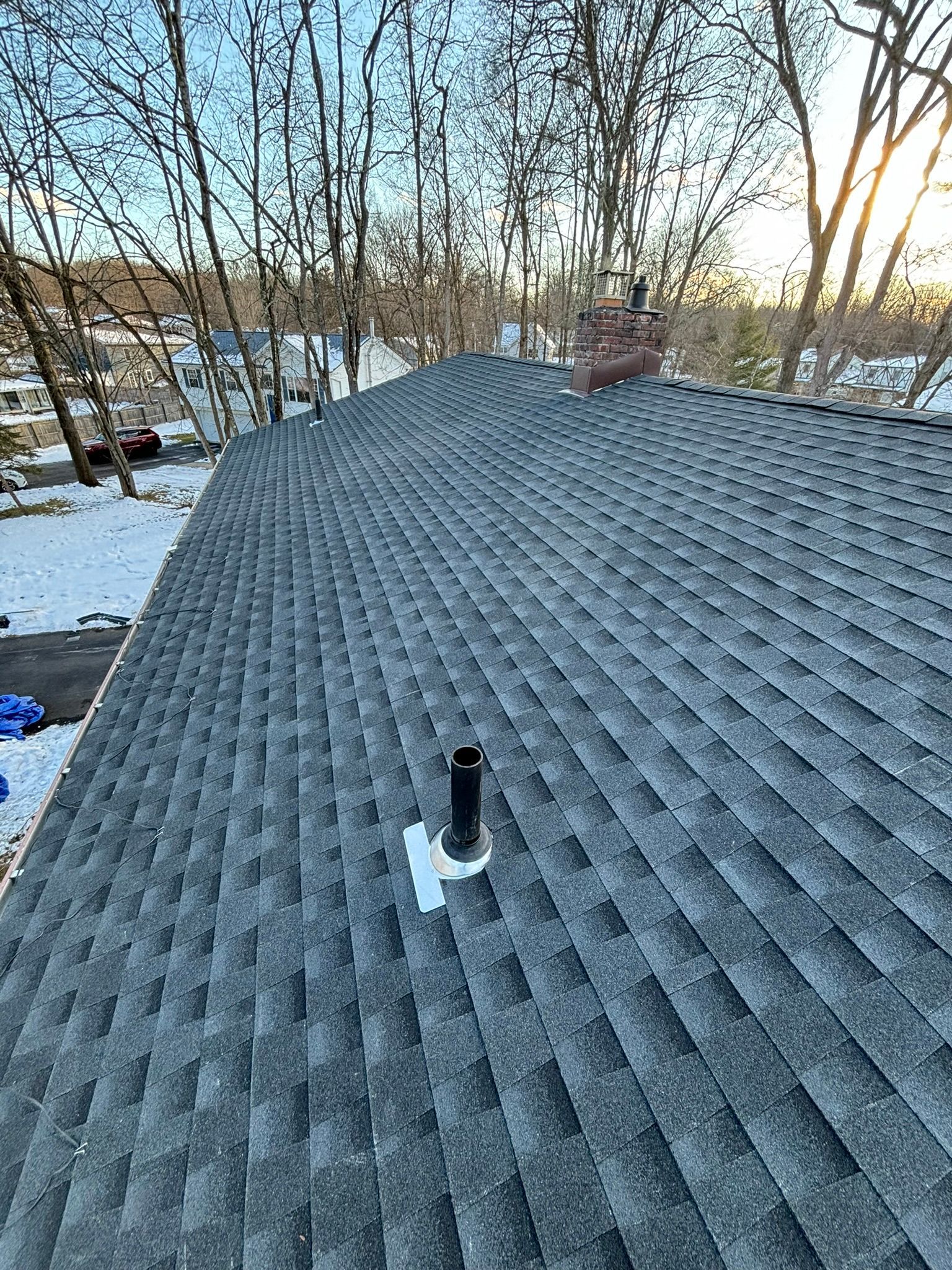 A gray shingle roof with a chimney and a pipe, in a snowy, wooded setting.