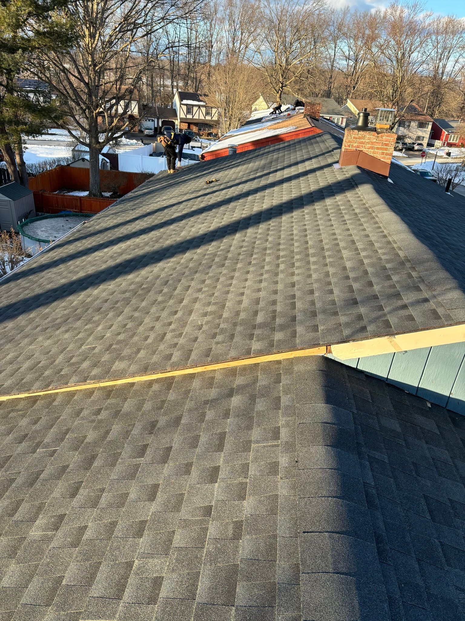 A rooftop with dark shingles, partially snow-covered, has workers and a chimney. The sky is clear.