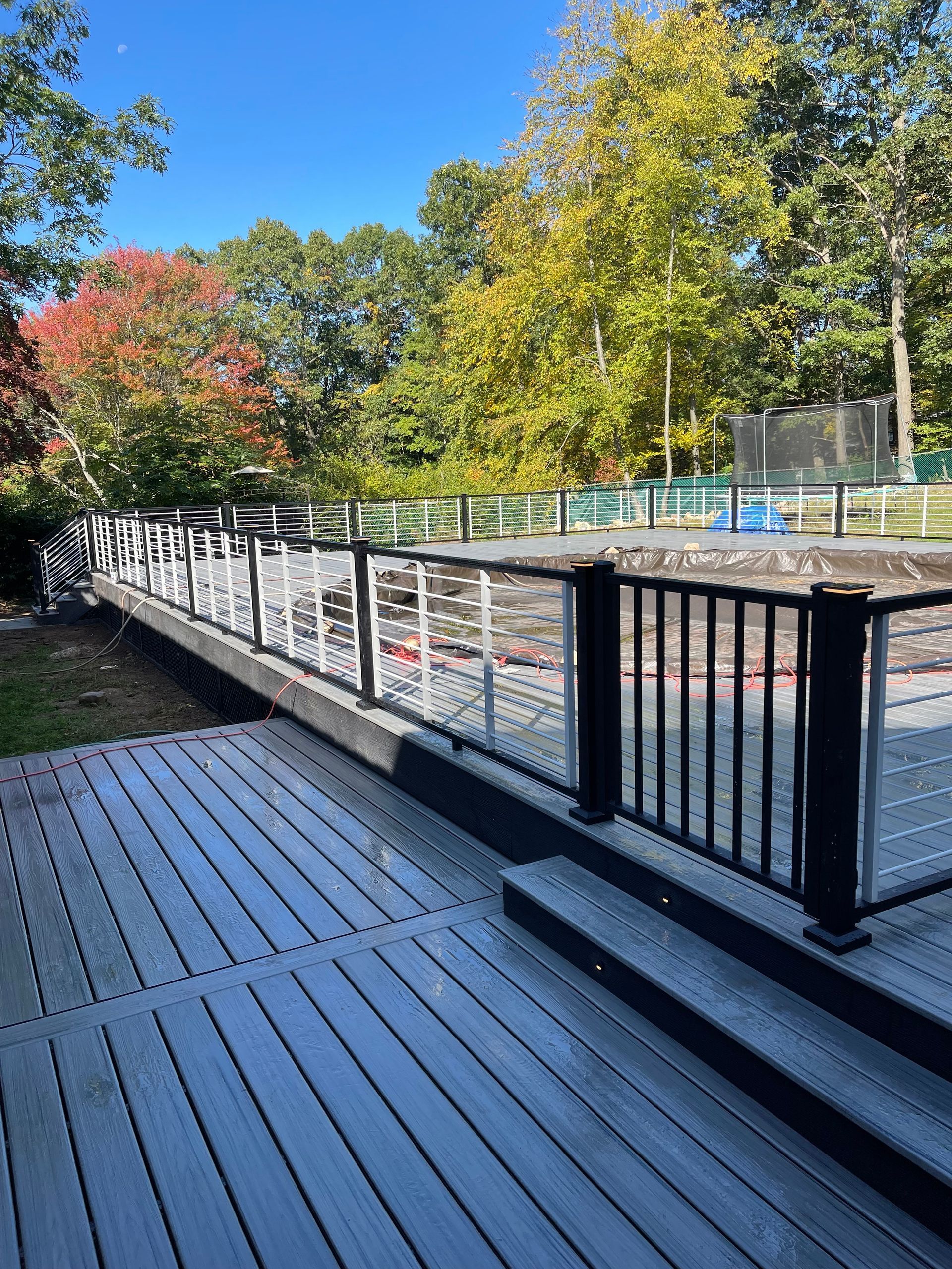 Wooden deck with black railing, leading to a smaller platform with white chairs and a black fence, trees in background.