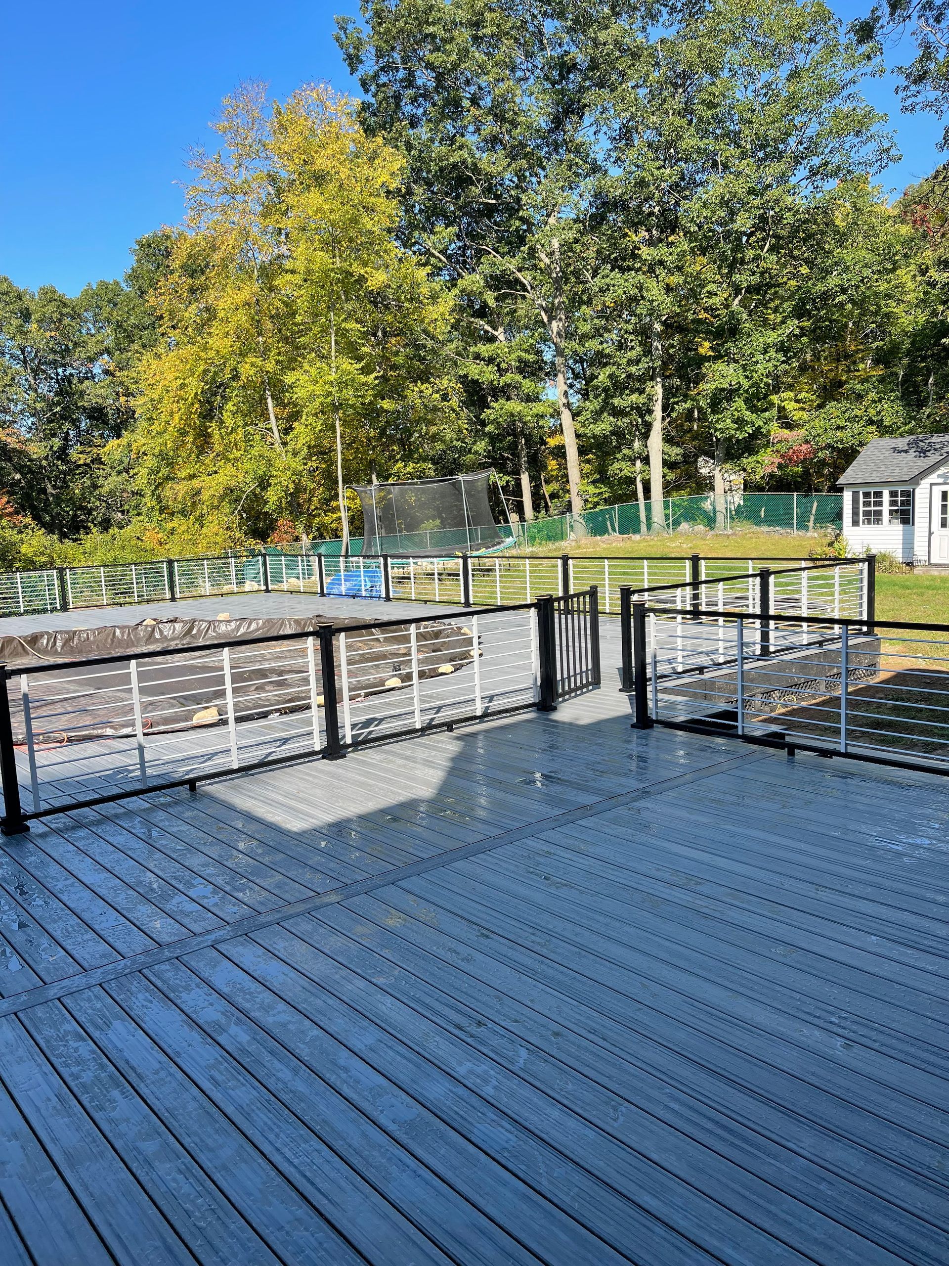 Deck overlooking a pool with a fence. Trees and a blue sky in the background.