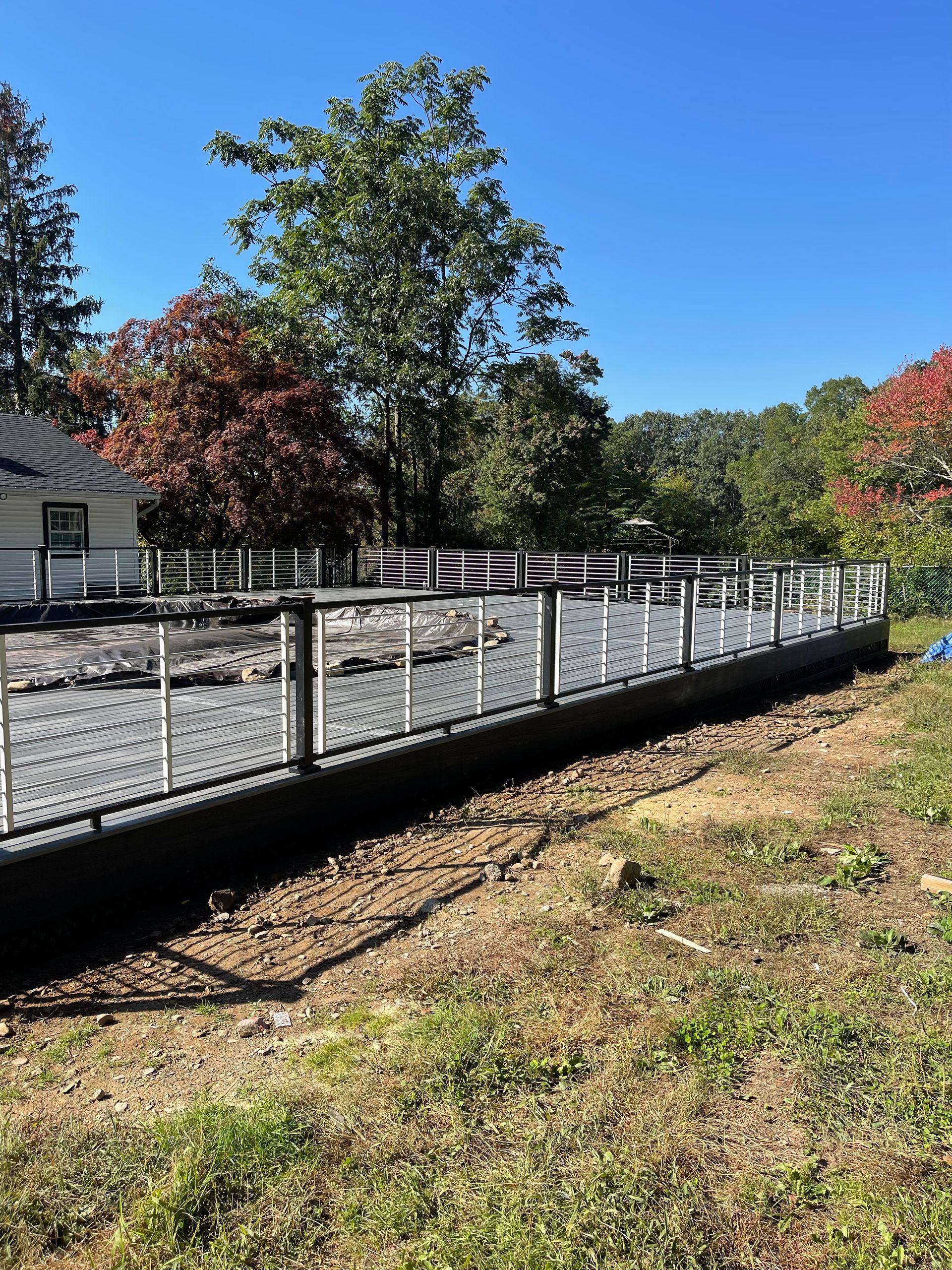 Gray and white fence along a dark, raised area with a house and trees in the background, under a blue sky.