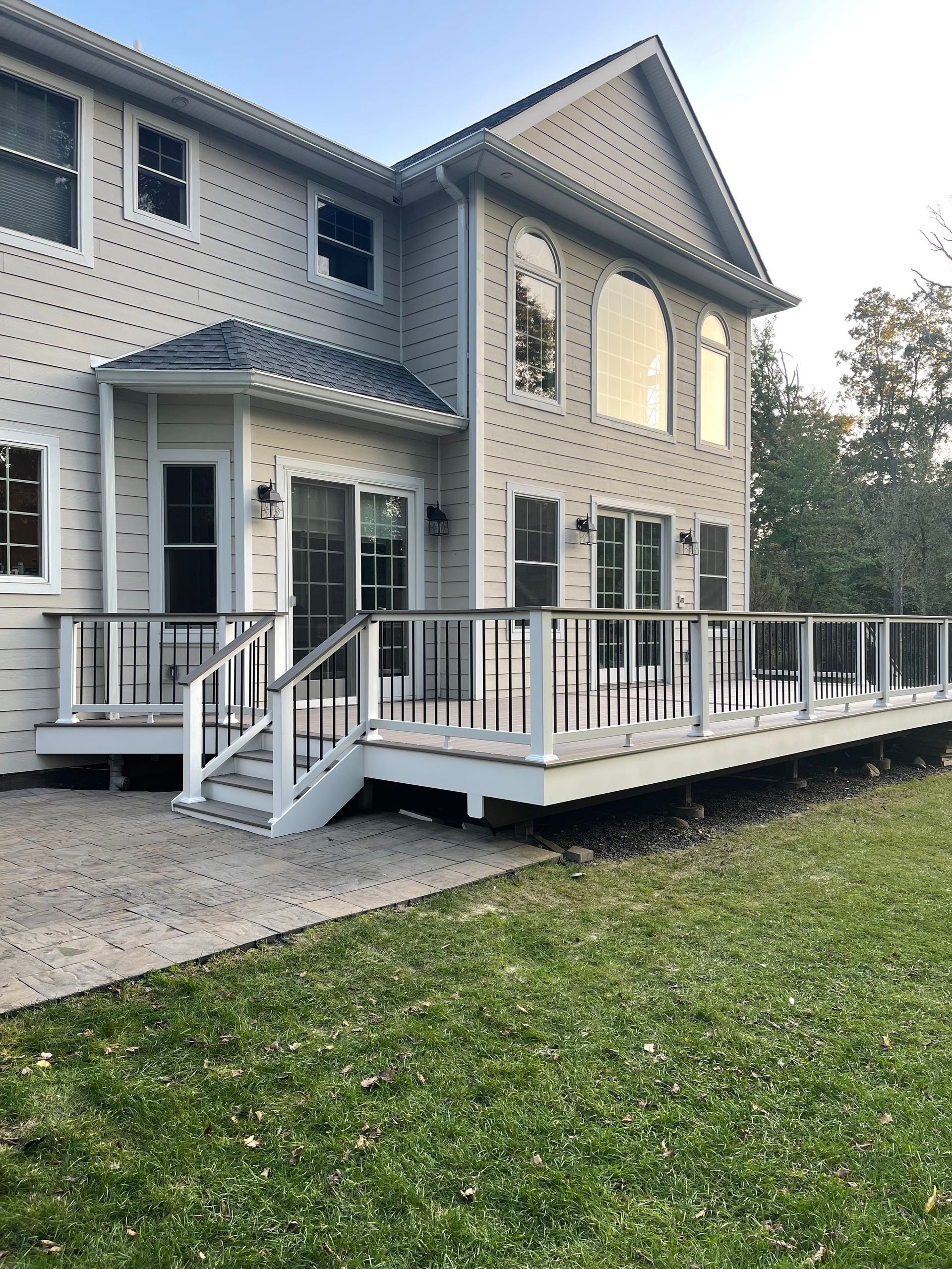 Back of a two-story beige house with a large wooden deck, black and white railing, and a grassy yard.