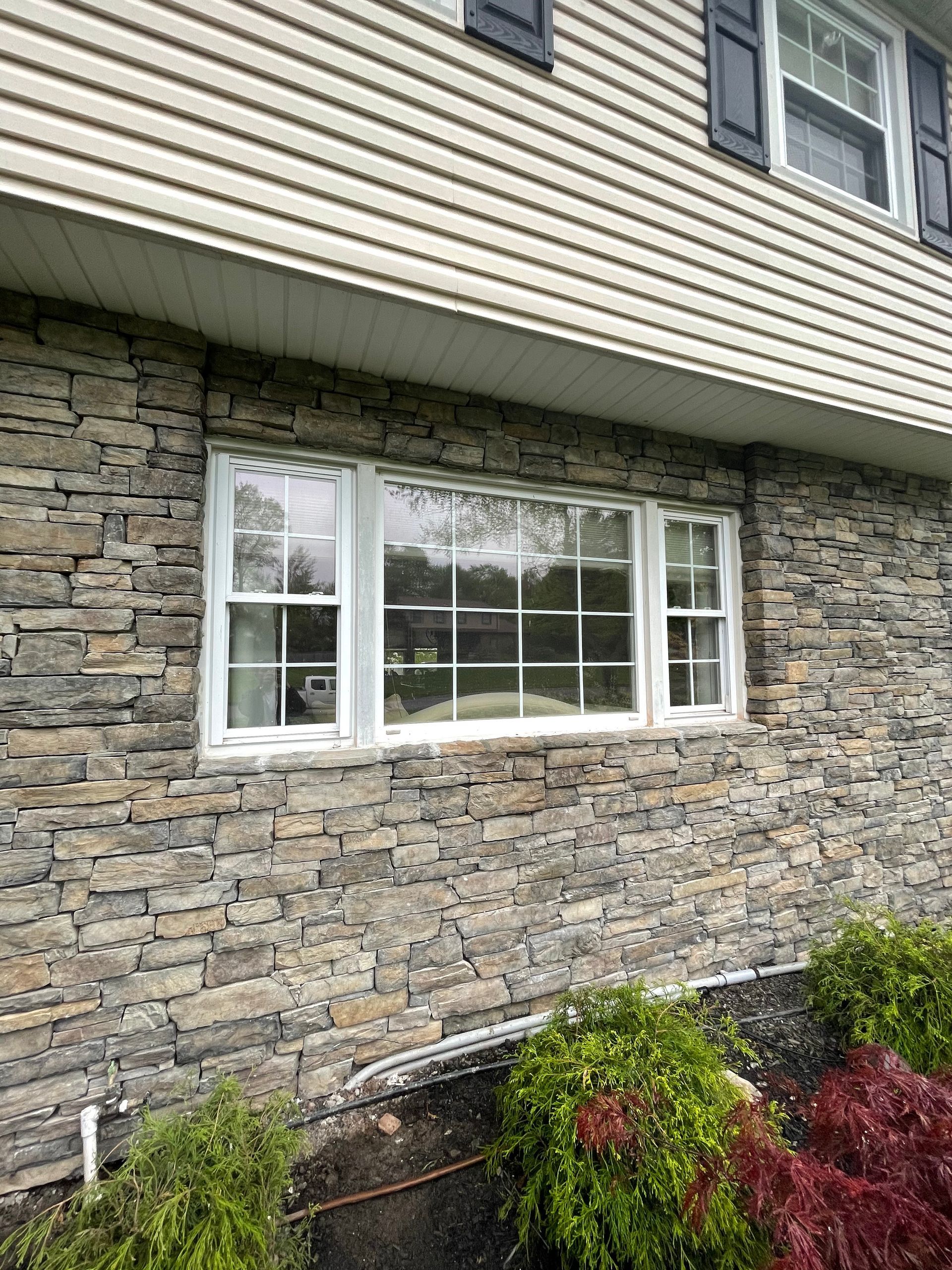 Stone-clad house with white-framed window. Dark shutters on upper level. Bushes at base.