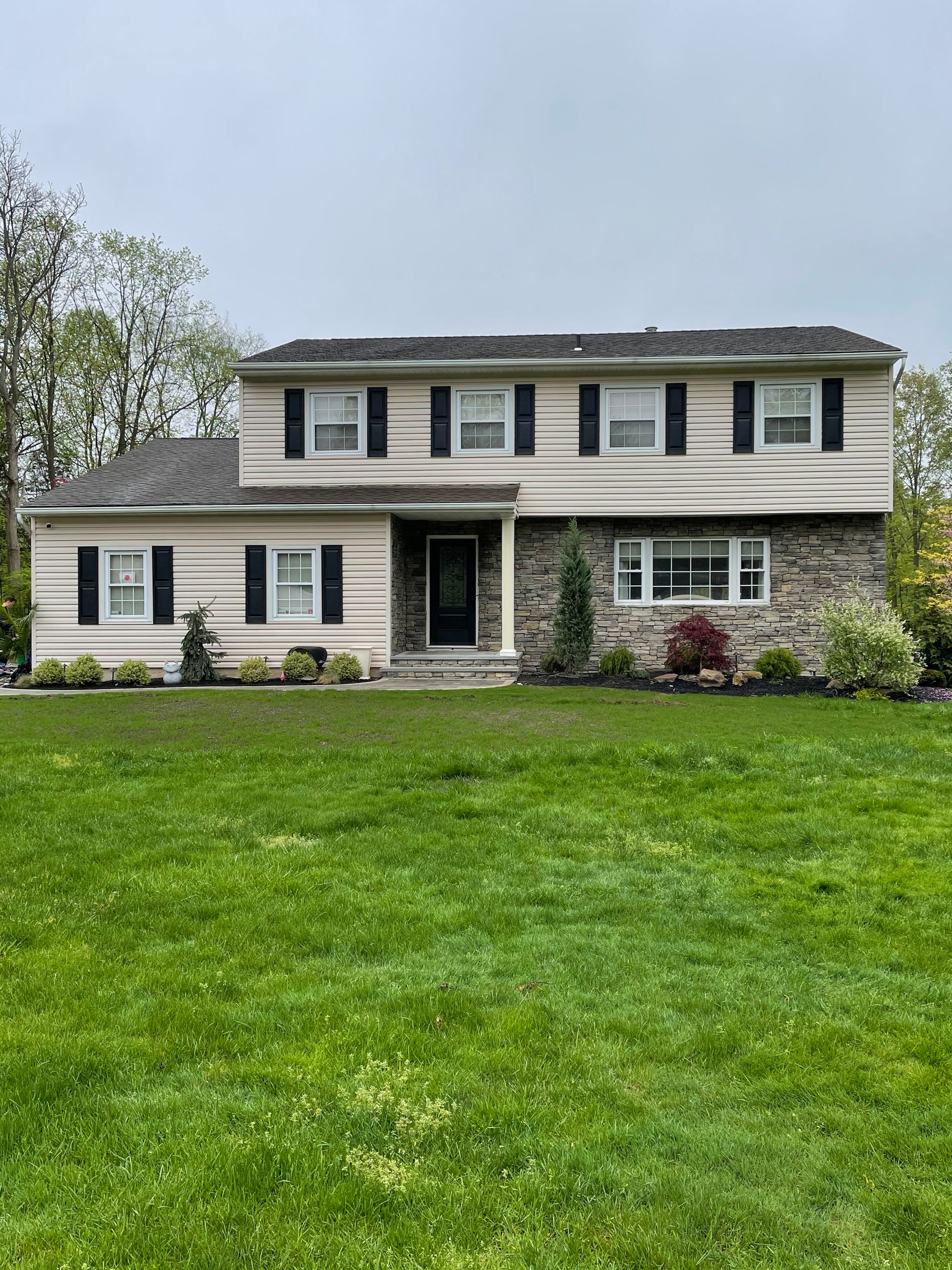 Two-story house with tan siding, stone accents, black shutters, and a green lawn.