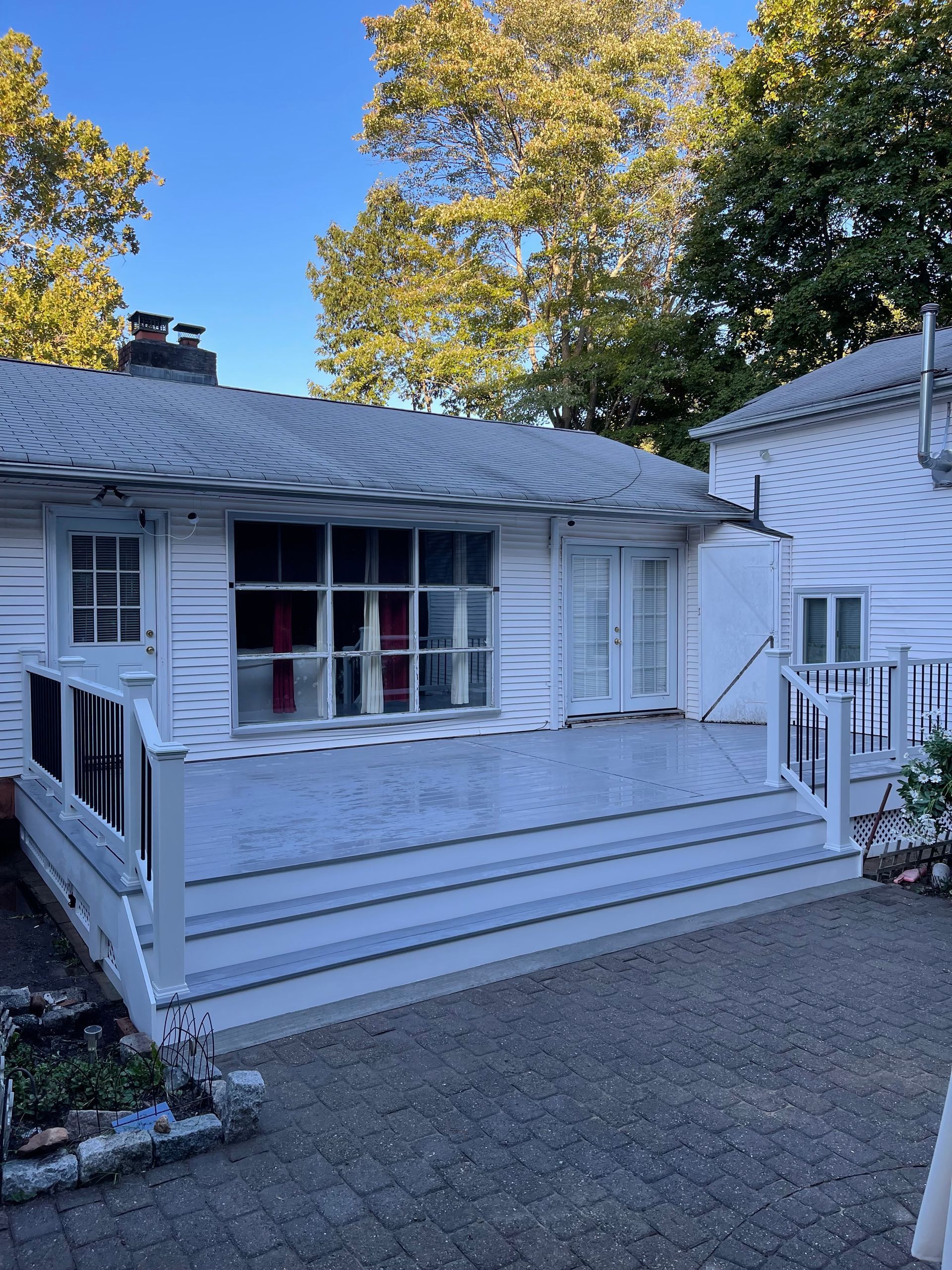 White house with porch and steps, gray patio. Trees and blue sky in background.