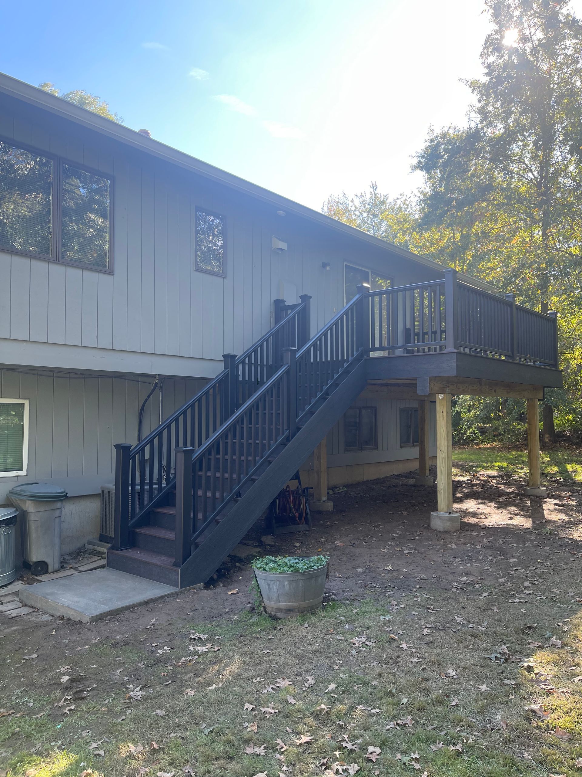 Gray house with black deck and stairs. Wooden railings.  Green plants, trees, and grass.