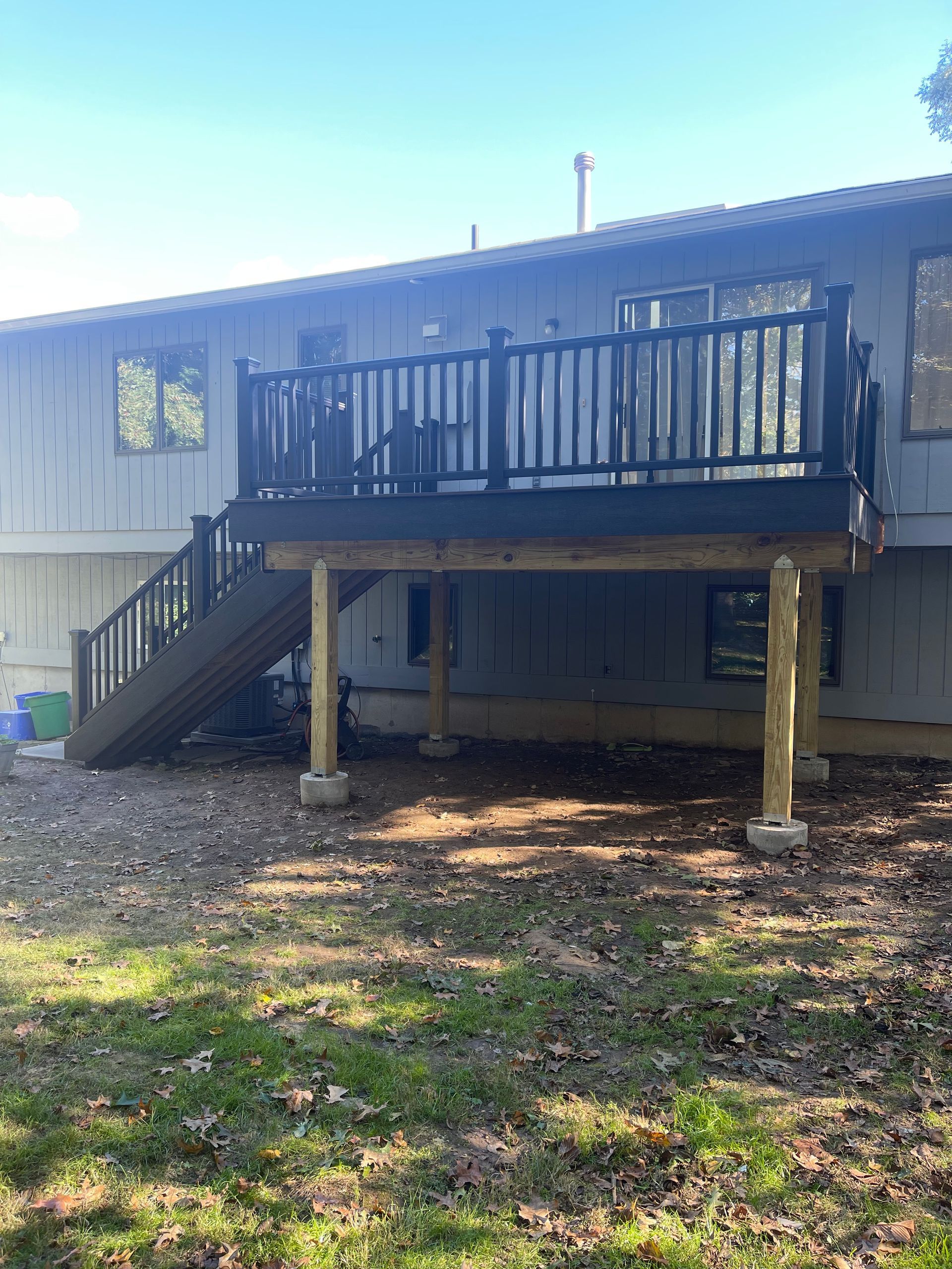 A two-story deck with black railing on a house. Wooden steps lead to the deck. The yard is covered with leaves.