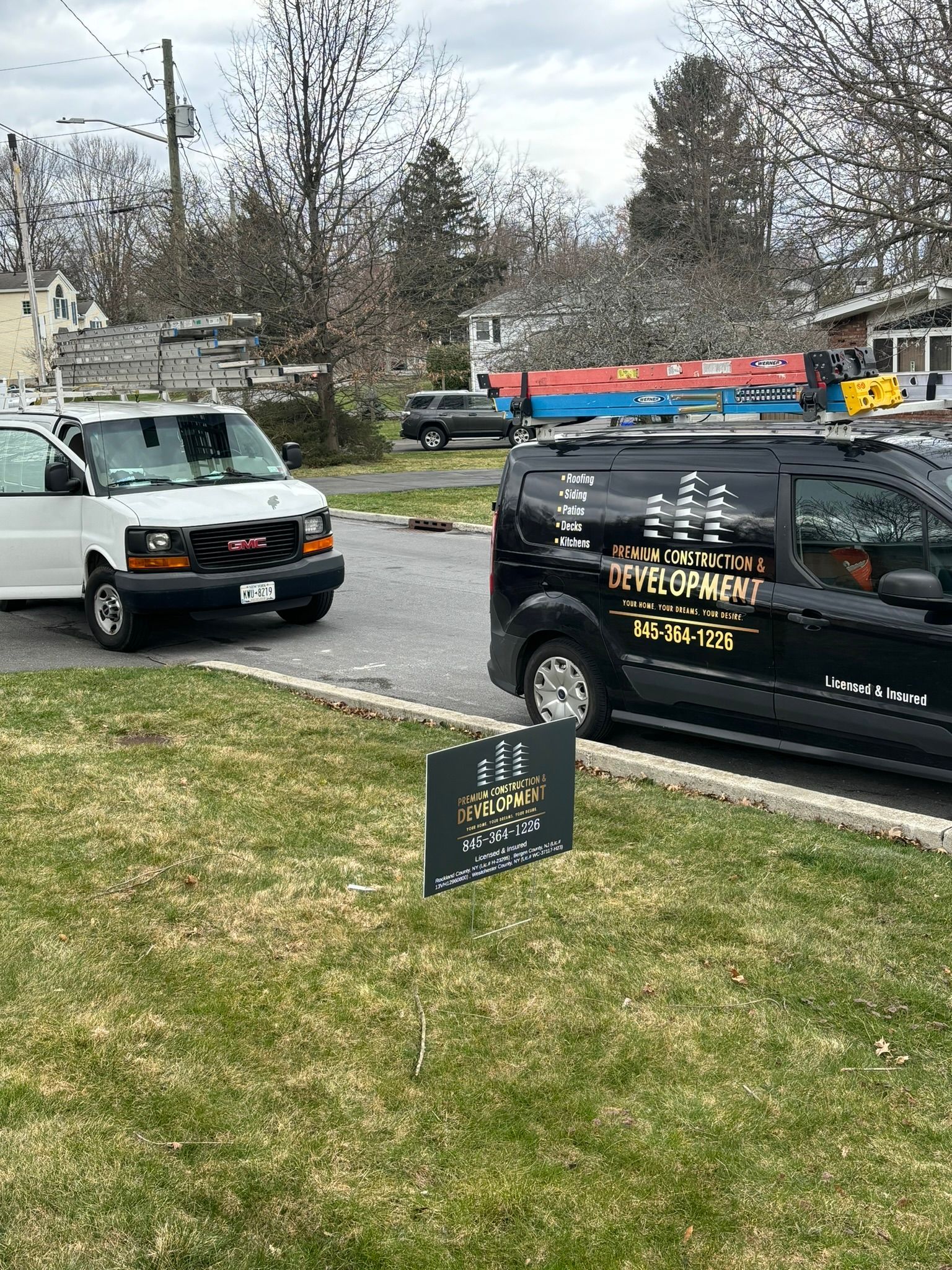 Two work vans parked on a residential street, yard sign in foreground.