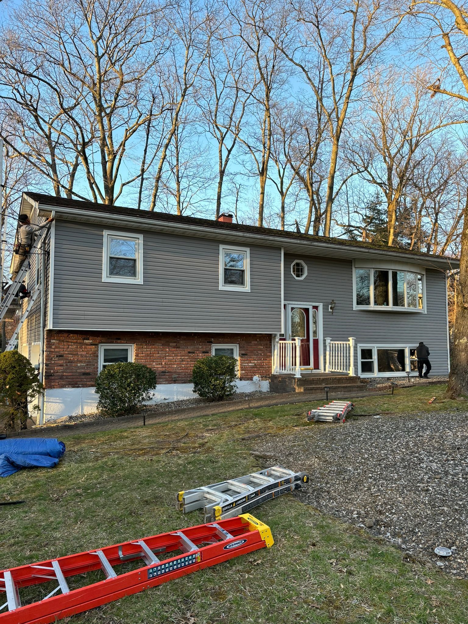 Two-story house with gray siding, red brick base, and a red ladder in the foreground.