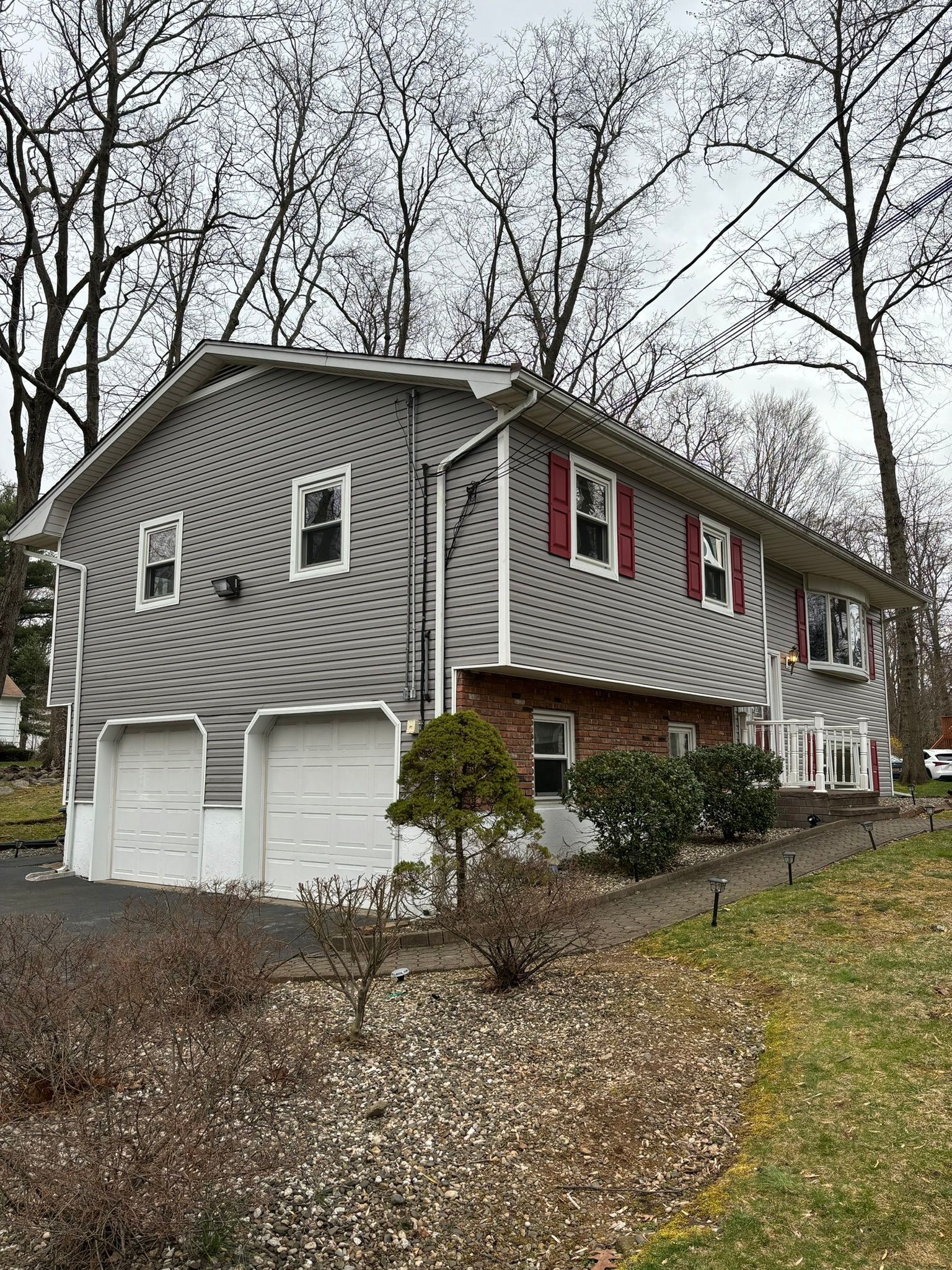 Two-story gray house with two-car garage, red shutters, and a brick section; surrounded by trees and landscaping.