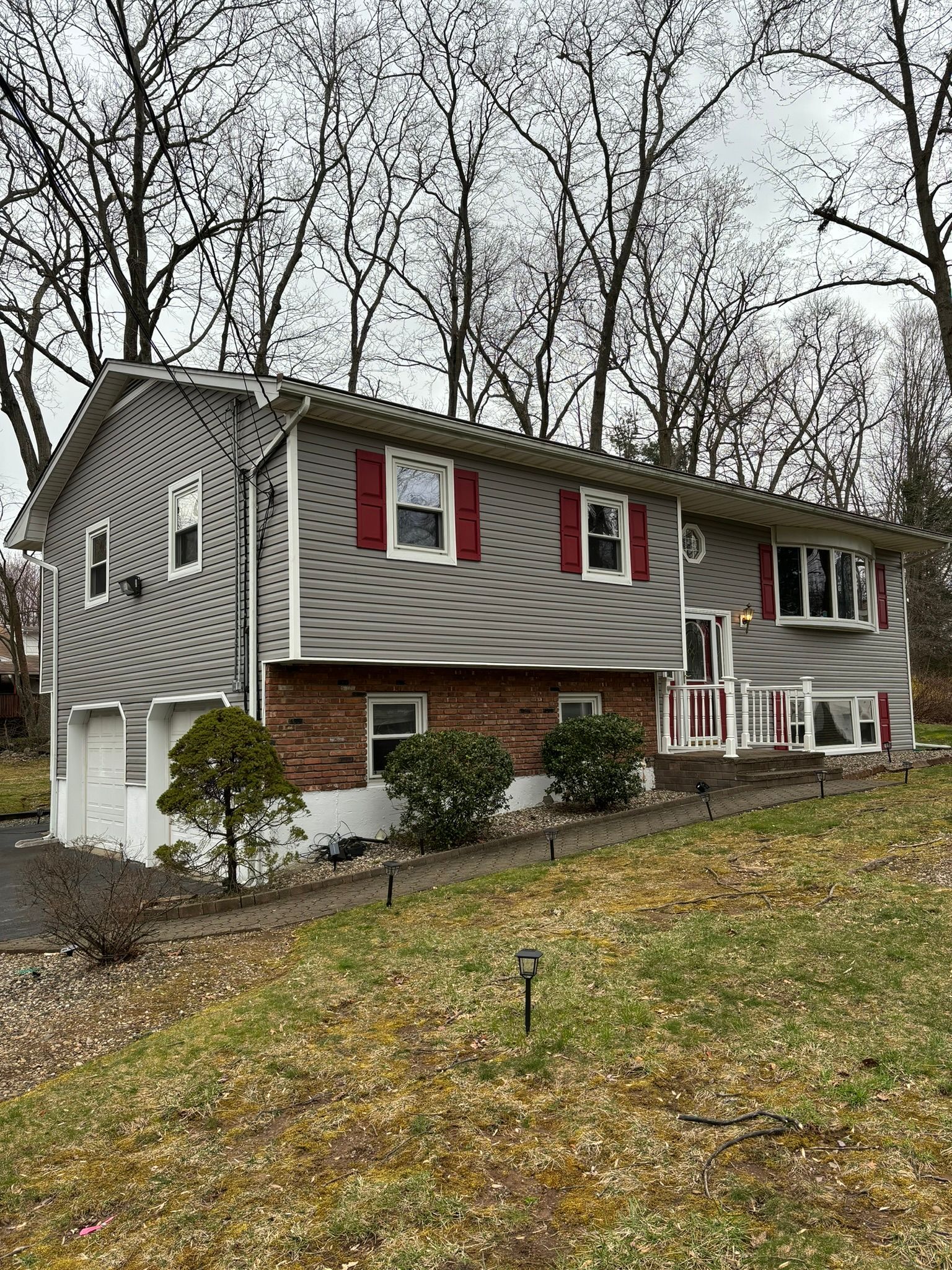 Two-story house with gray siding, red shutters, brick facade, and a sloped lawn.