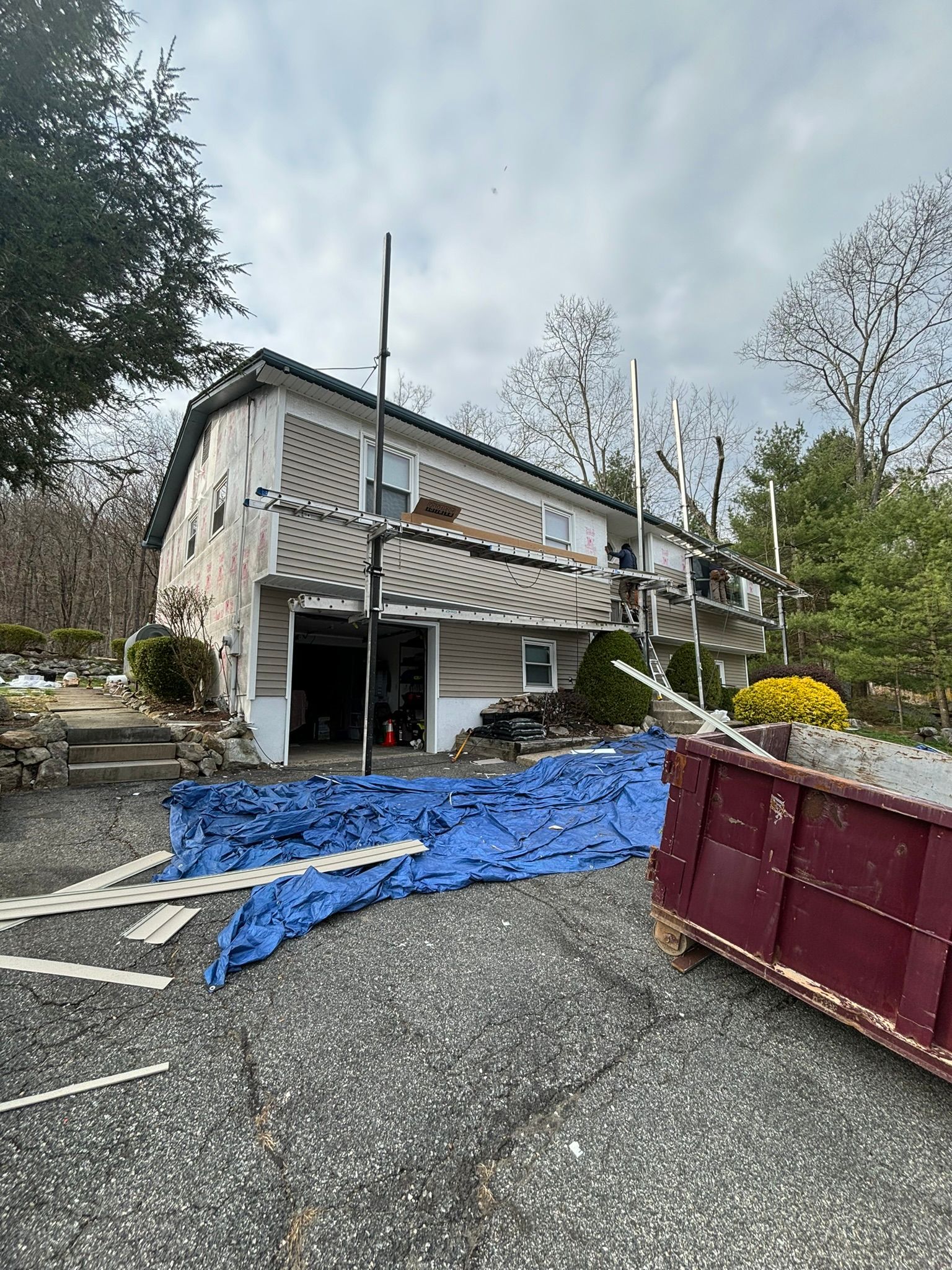 House under construction, scaffolding, debris. Blue tarp in driveway, dumpster. Gray, beige, and cloudy sky.
