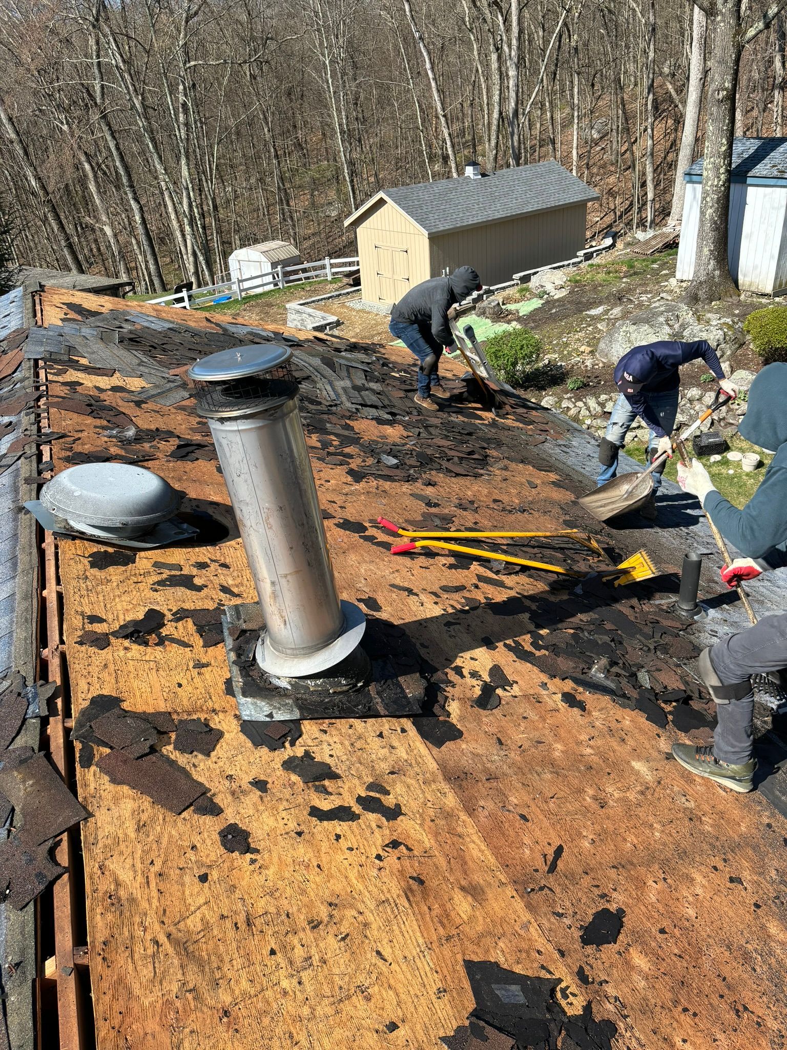 People on a roof removing old shingles, chimney visible, sunny day, trees in the background.