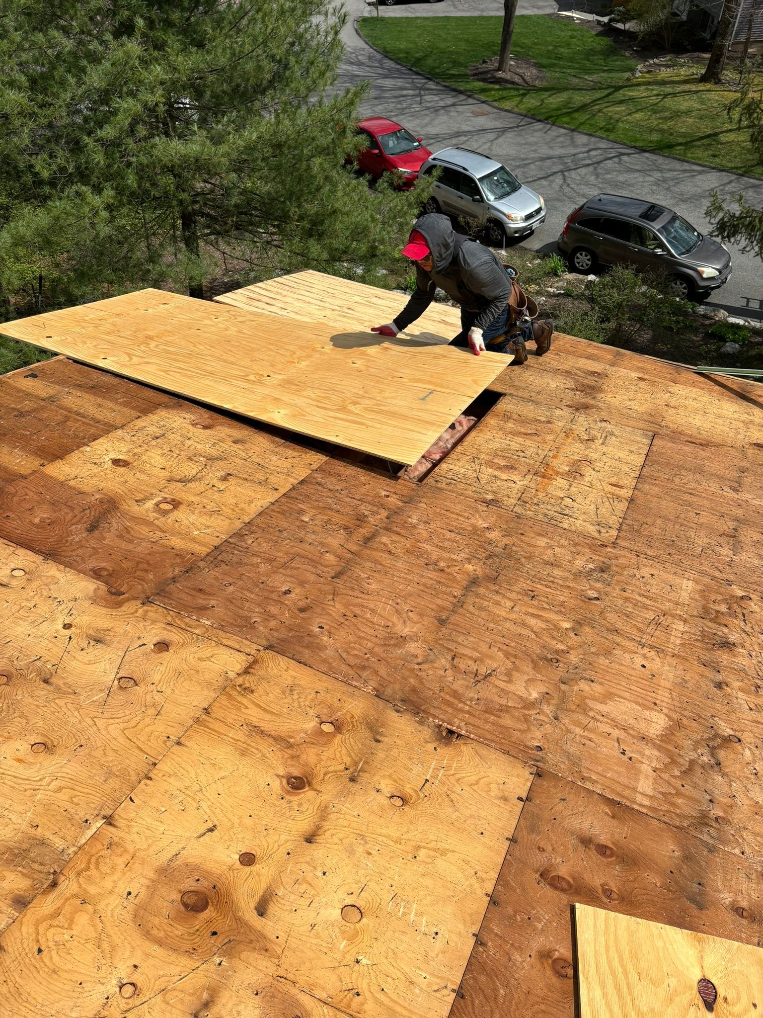 Two workers install plywood on a roof. Cars parked on a street are visible in the background.