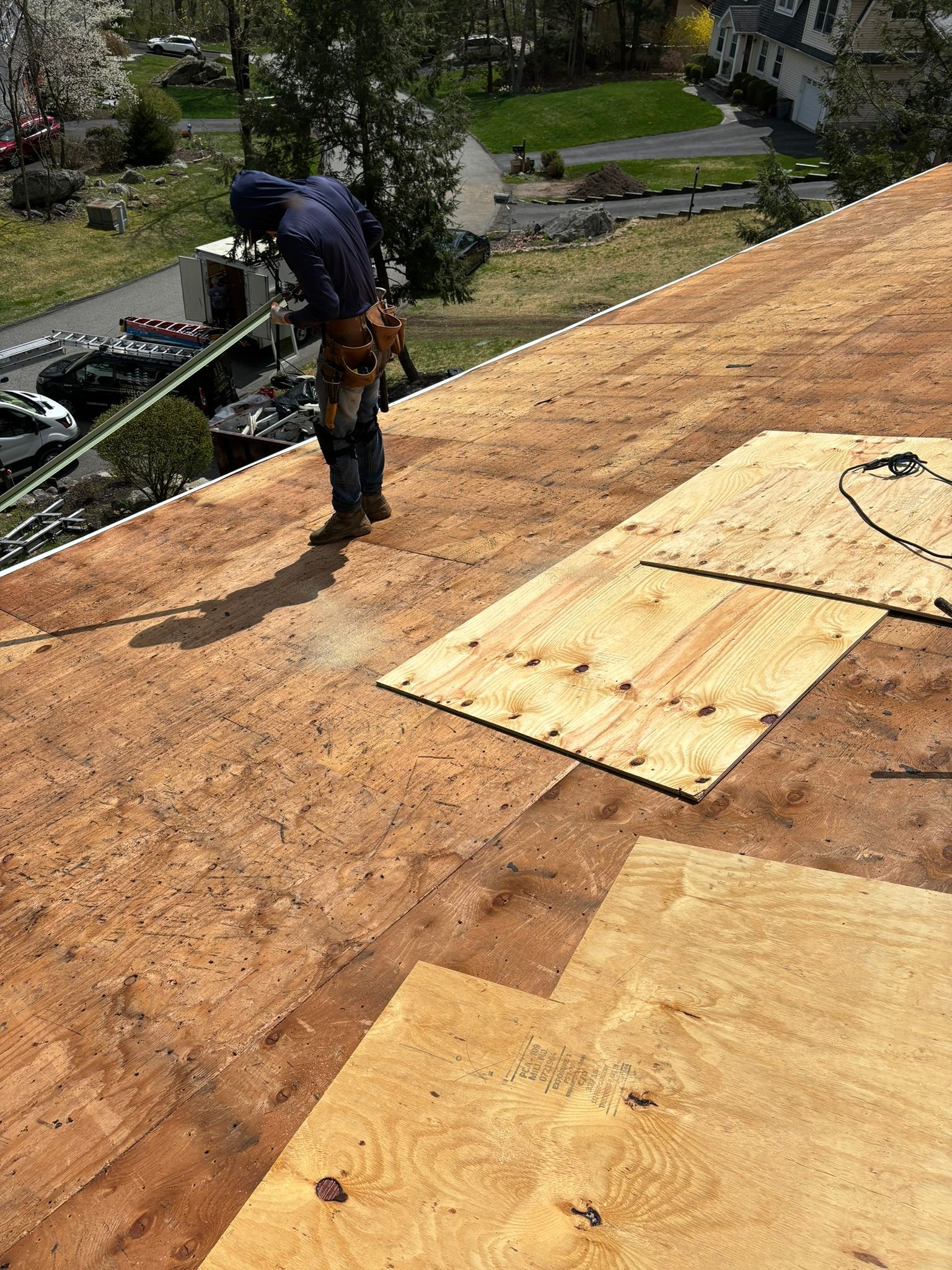 Roofer working on a roof, surrounded by plywood sheets, in front of a residential neighborhood.