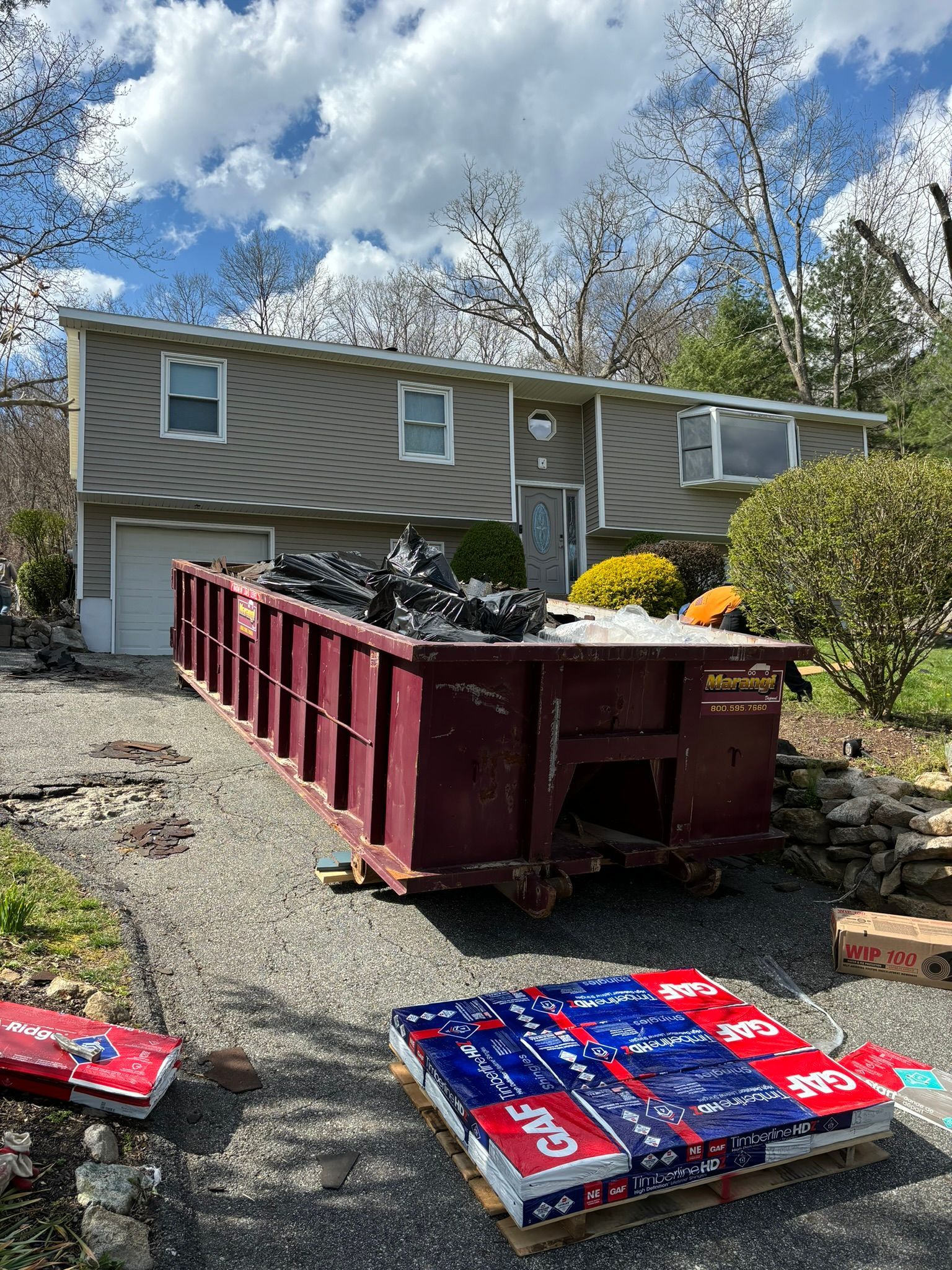 A dumpster sits in front of a house with stacked roofing tiles on the ground.