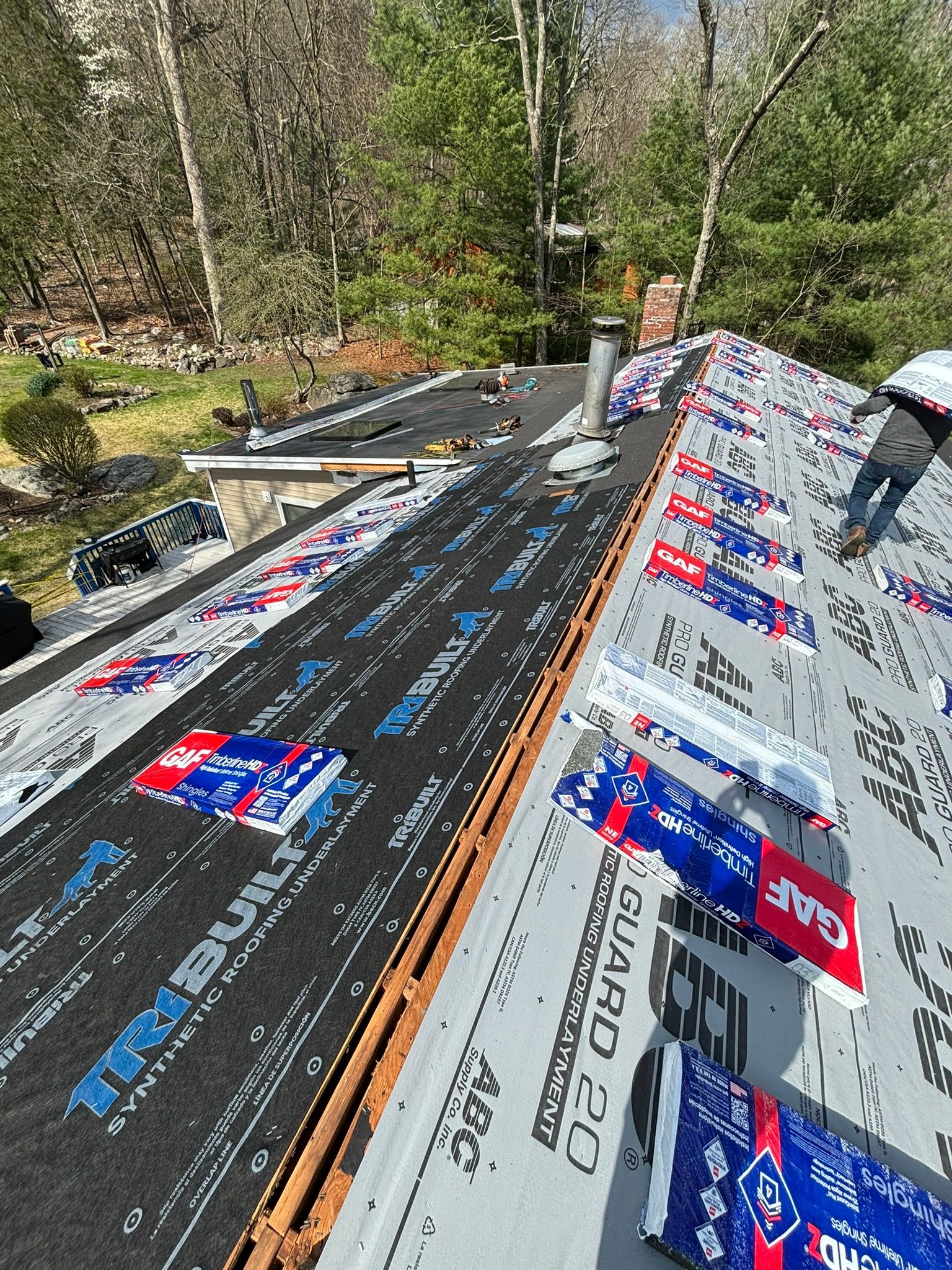 Workers installing roofing material on a house roof, surrounded by trees.