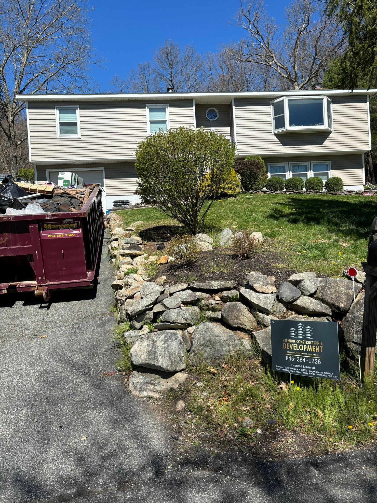 House with a stone retaining wall, driveway with dumpster, and a small sign on the front lawn.