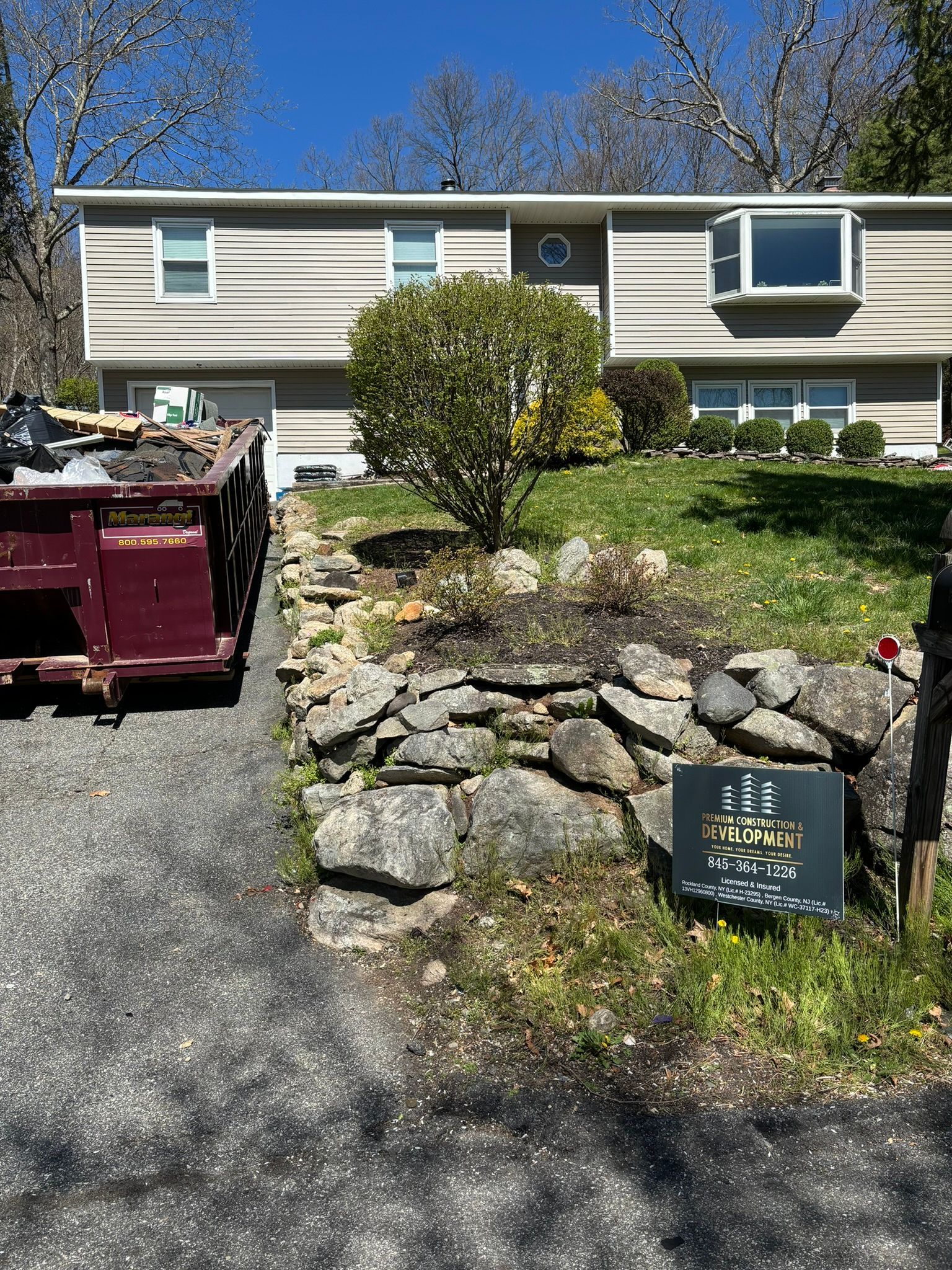 Two-story house with a dark dumpster in the driveway and a stone wall in front.